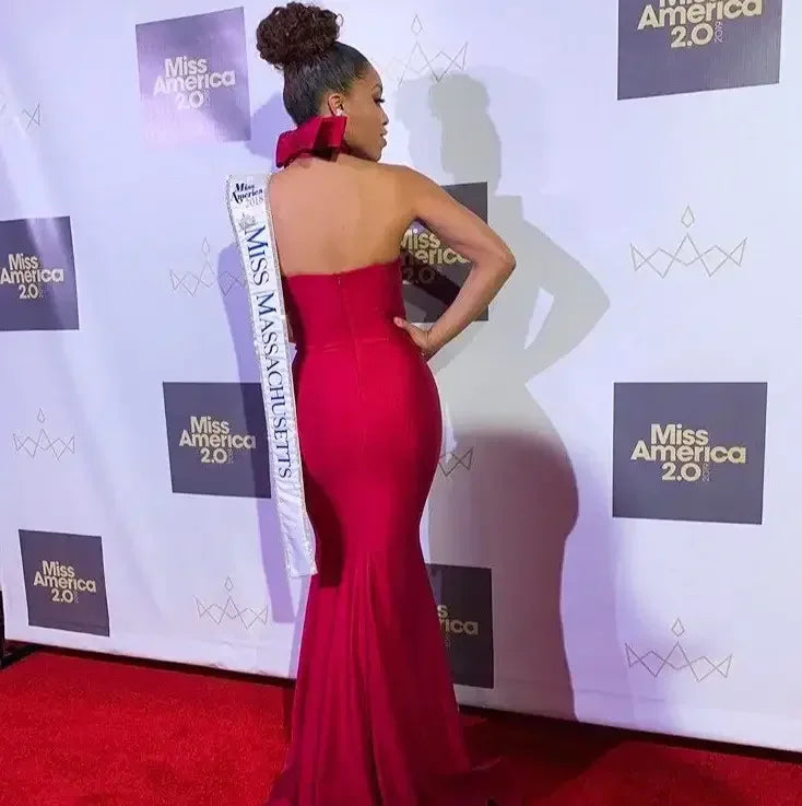 A woman in a fitted red gown stands confidently, wearing a sash that reads "Miss Massachusetts," against a backdrop displaying "Miss America 2.0" on a red carpet event.