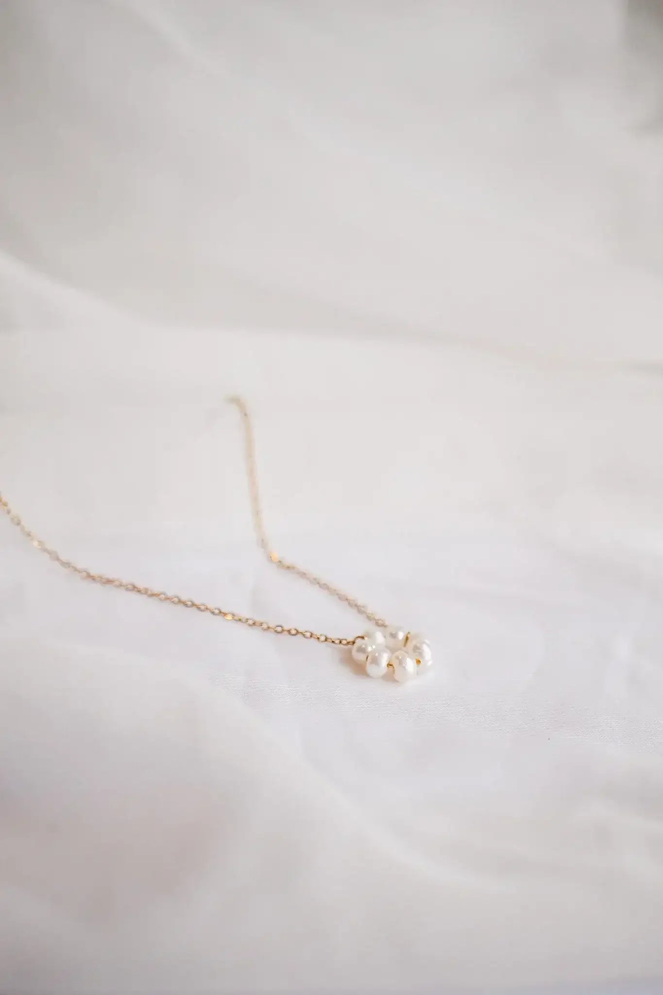 A delicate gold necklace with a cluster of white pearls rests on soft white fabric, highlighting its elegant design in a minimalistic setting.