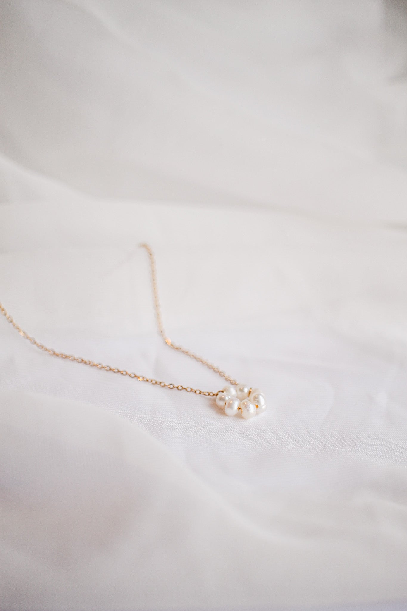 A delicate gold necklace with a cluster of white pearls rests on soft white fabric, highlighting its elegant design in a minimalistic setting.