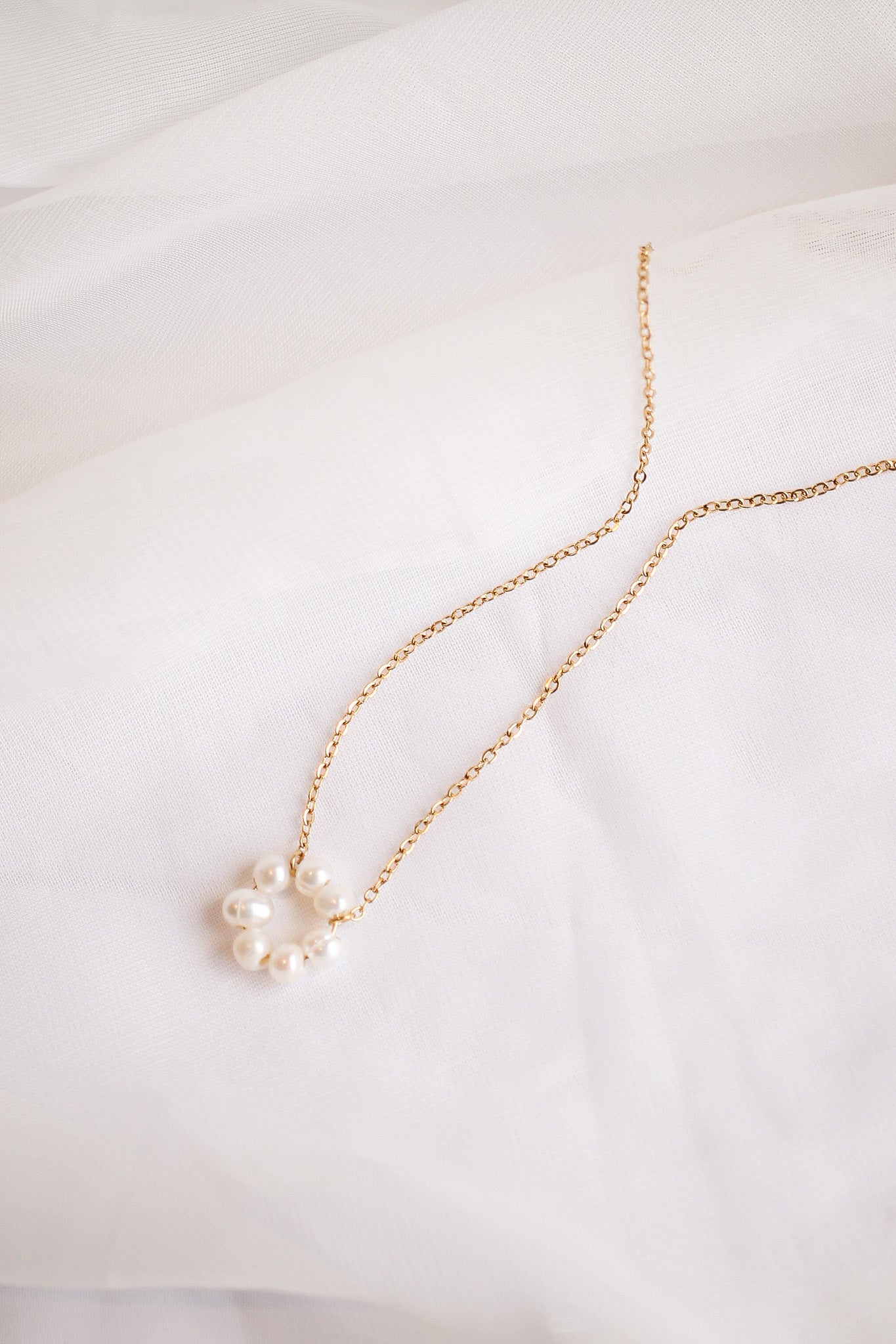 A delicate gold necklace with a cluster of small pearls rests on soft, white fabric. The pearls are arranged in a circular pattern, enhancing the necklace's elegance.