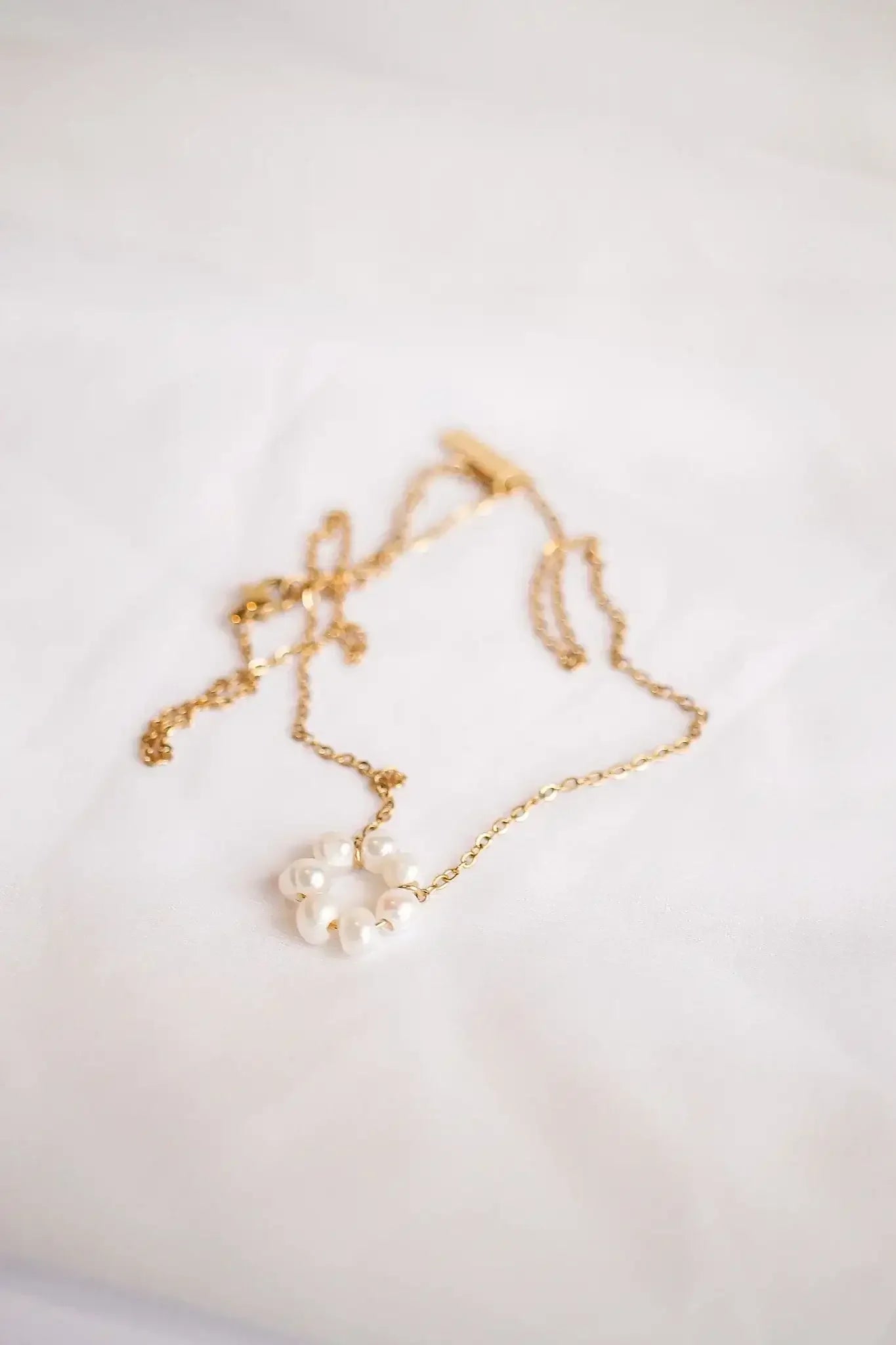 A delicate gold necklace with a circular cluster of white pearls rests on a soft, white fabric background, reflecting an elegant and sophisticated aesthetic.