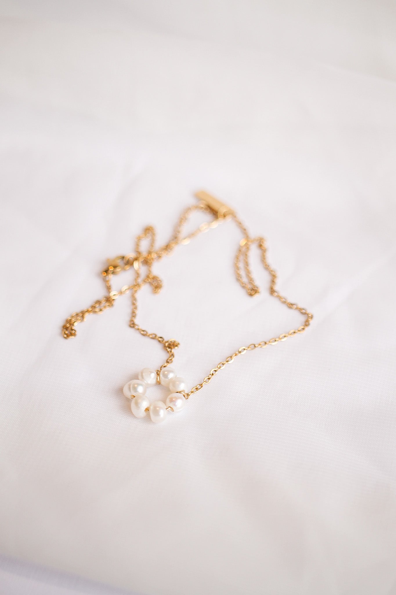 A delicate gold necklace with a circular cluster of white pearls rests on a soft, white fabric background, reflecting an elegant and sophisticated aesthetic.