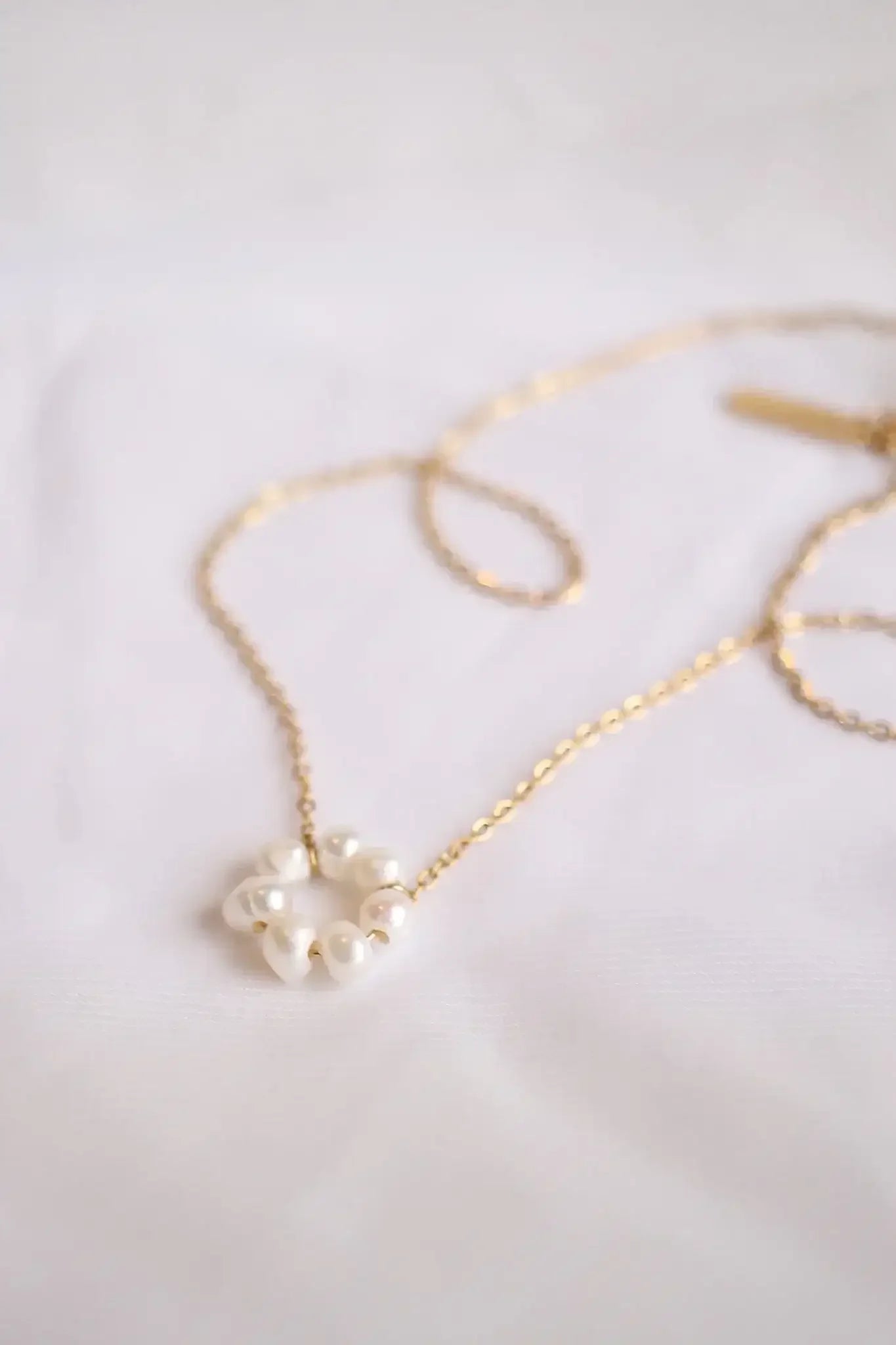 A delicate gold necklace features a circular arrangement of white pearls at its center, resting on a soft white fabric background, showcasing an elegant and minimalistic design.