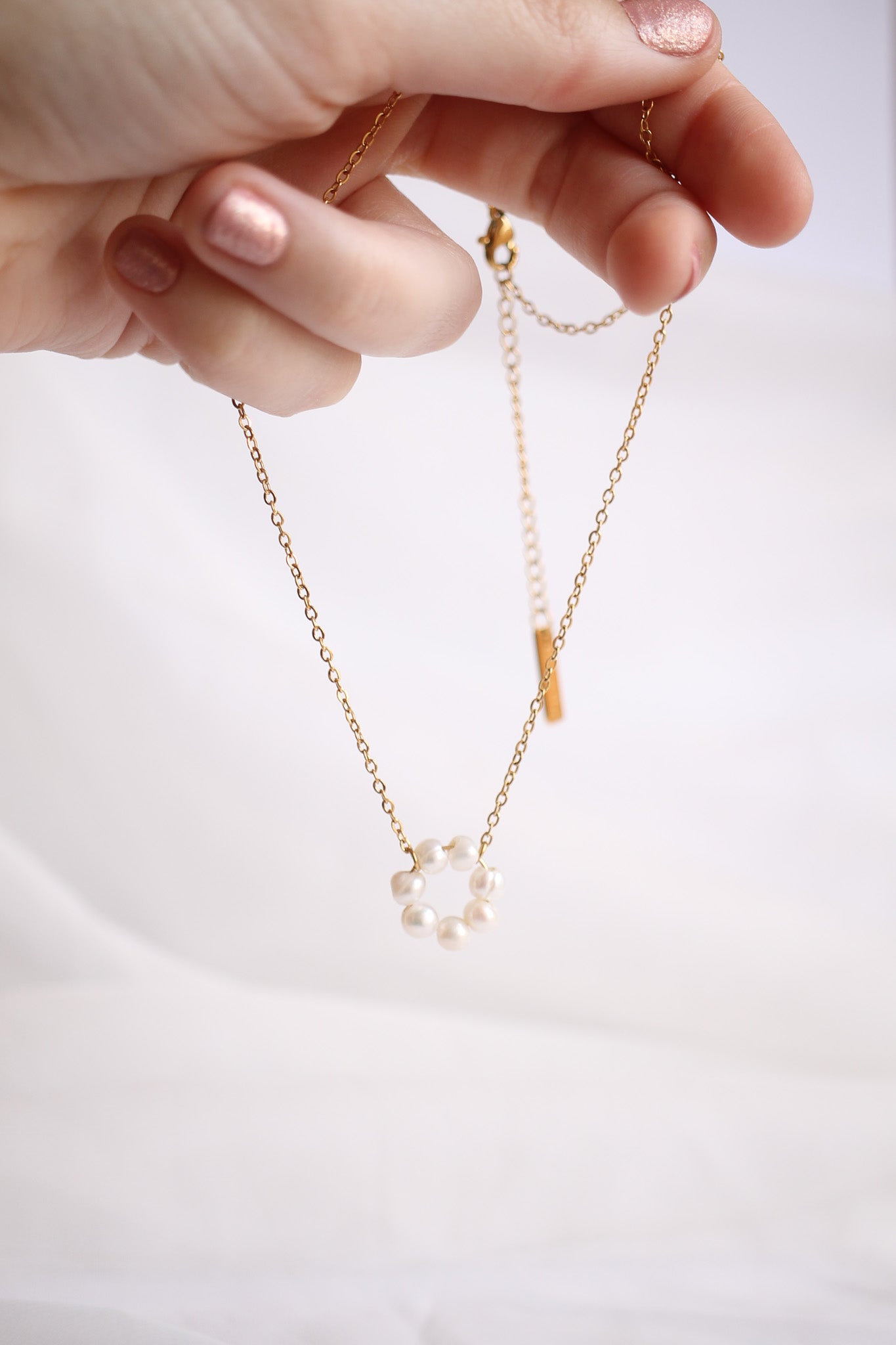 A hand holds a delicate gold chain necklace with a circular pearl pendant while set against a soft, light background. The pearls form a subtle, elegant design.