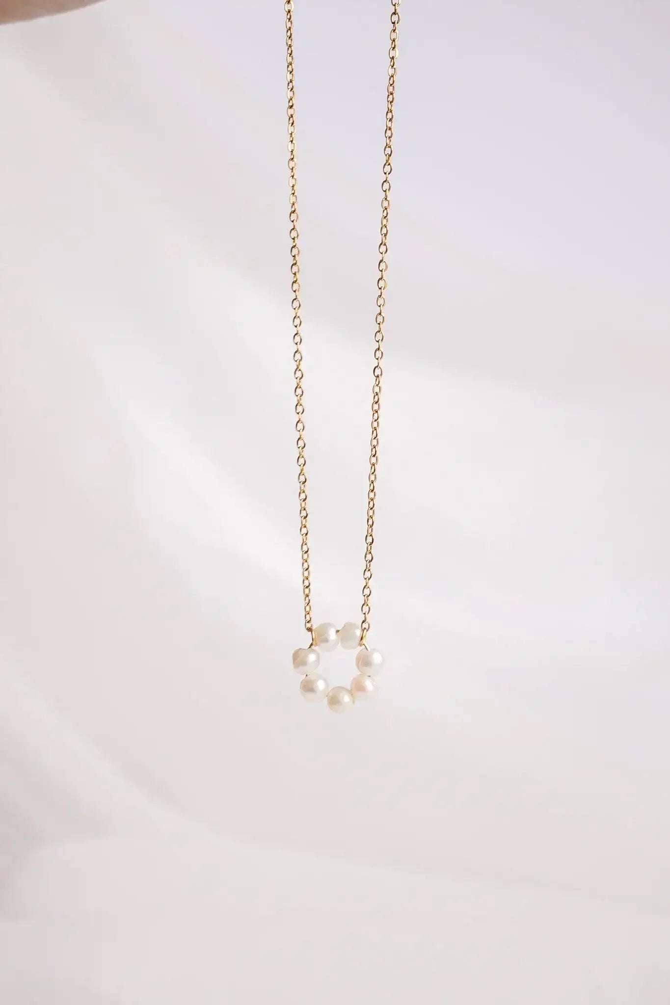 A gold necklace features a circular arrangement of small pearls hanging from a delicate chain, set against a soft, cream-colored background that enhances its elegance and simplicity.
