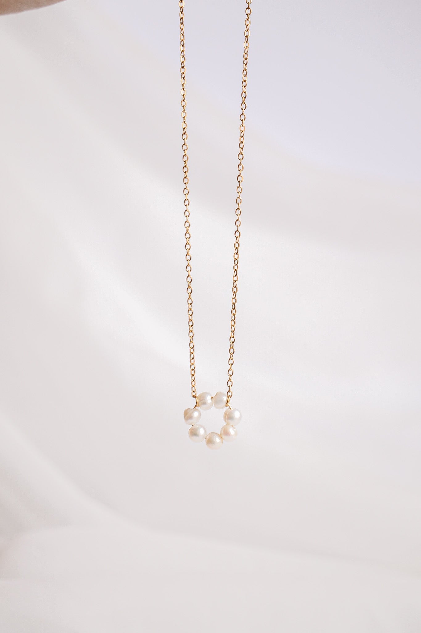 A gold necklace features a circular arrangement of small pearls hanging from a delicate chain, set against a soft, cream-colored background that enhances its elegance and simplicity.