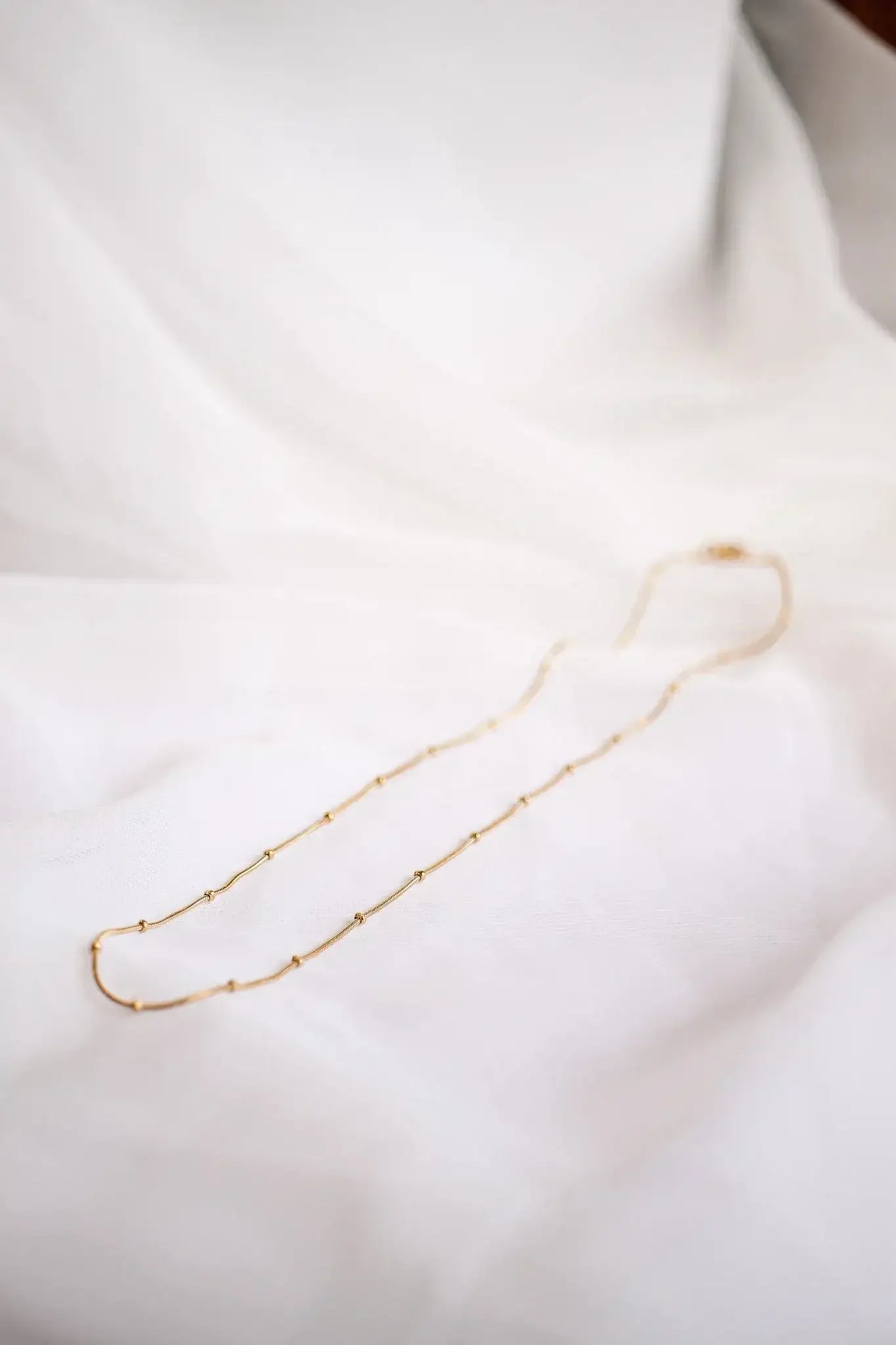 A delicate gold necklace lies elegantly on soft white fabric, showcasing its intertwined design and spaced beads, emphasizing its artistic craftsmanship in a serene, minimalist setting.
