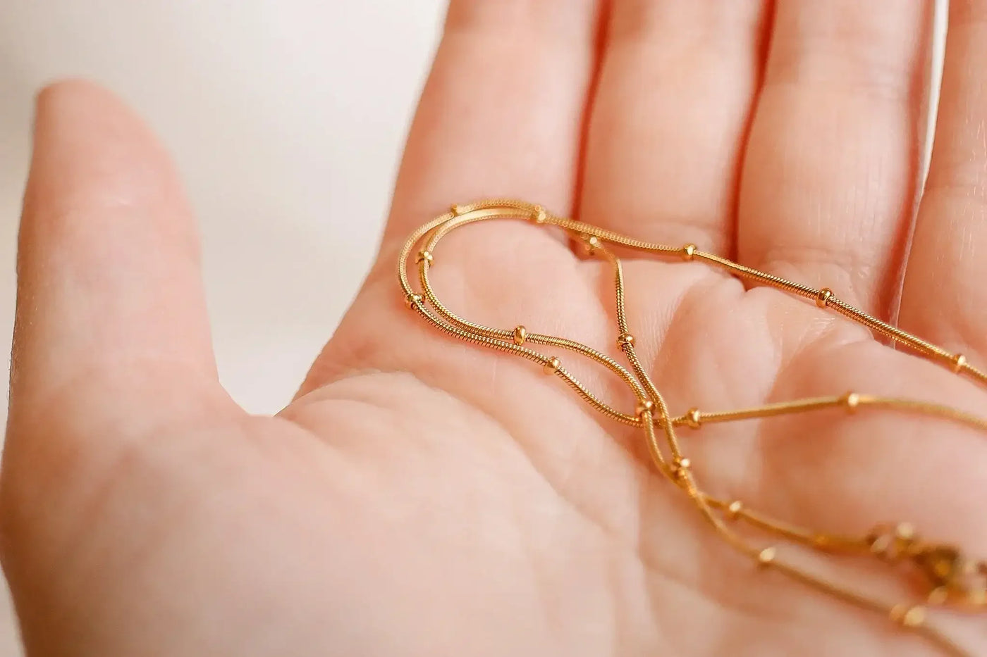 Delicate gold chains rest on an open palm, intertwining gracefully. The background is softly blurred, emphasizing the shine and intricate design of the jewelry against the skin.