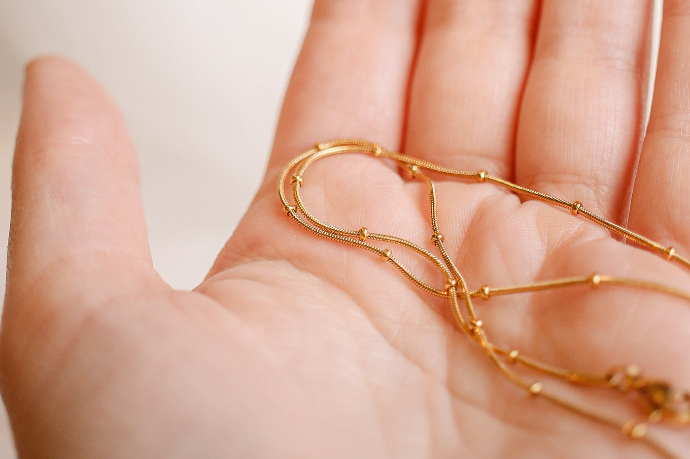 Delicate gold chains rest on an open palm, intertwining gracefully. The background is softly blurred, emphasizing the shine and intricate design of the jewelry against the skin.