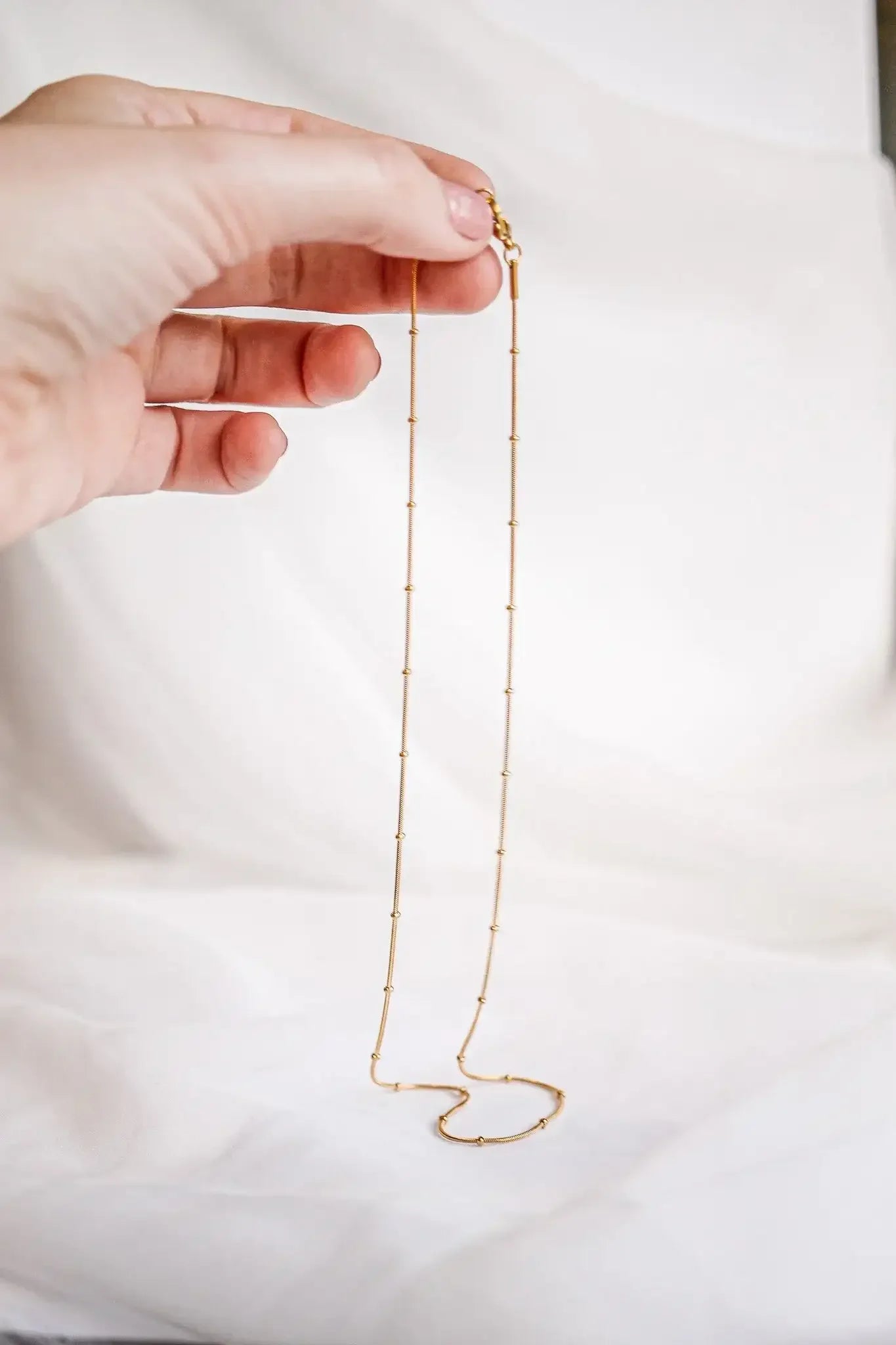 A hand holds a delicate gold necklace, showcasing its long, thin chain adorned with small round beads. The background features soft, white fabric, enhancing the necklace's elegance.