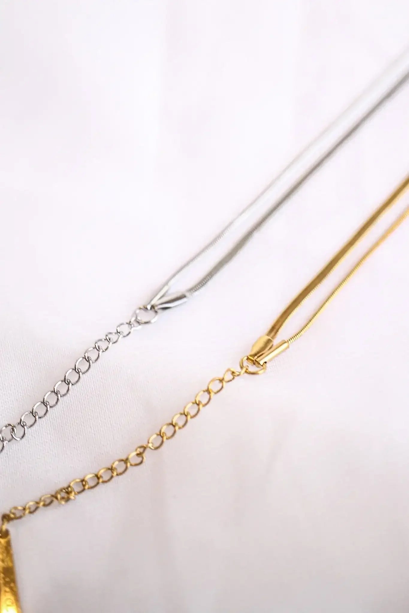 Two necklaces, one silver and one gold, lie flat against a soft, white background, displaying their chains intertwined with clasps positioned for potential fastening.