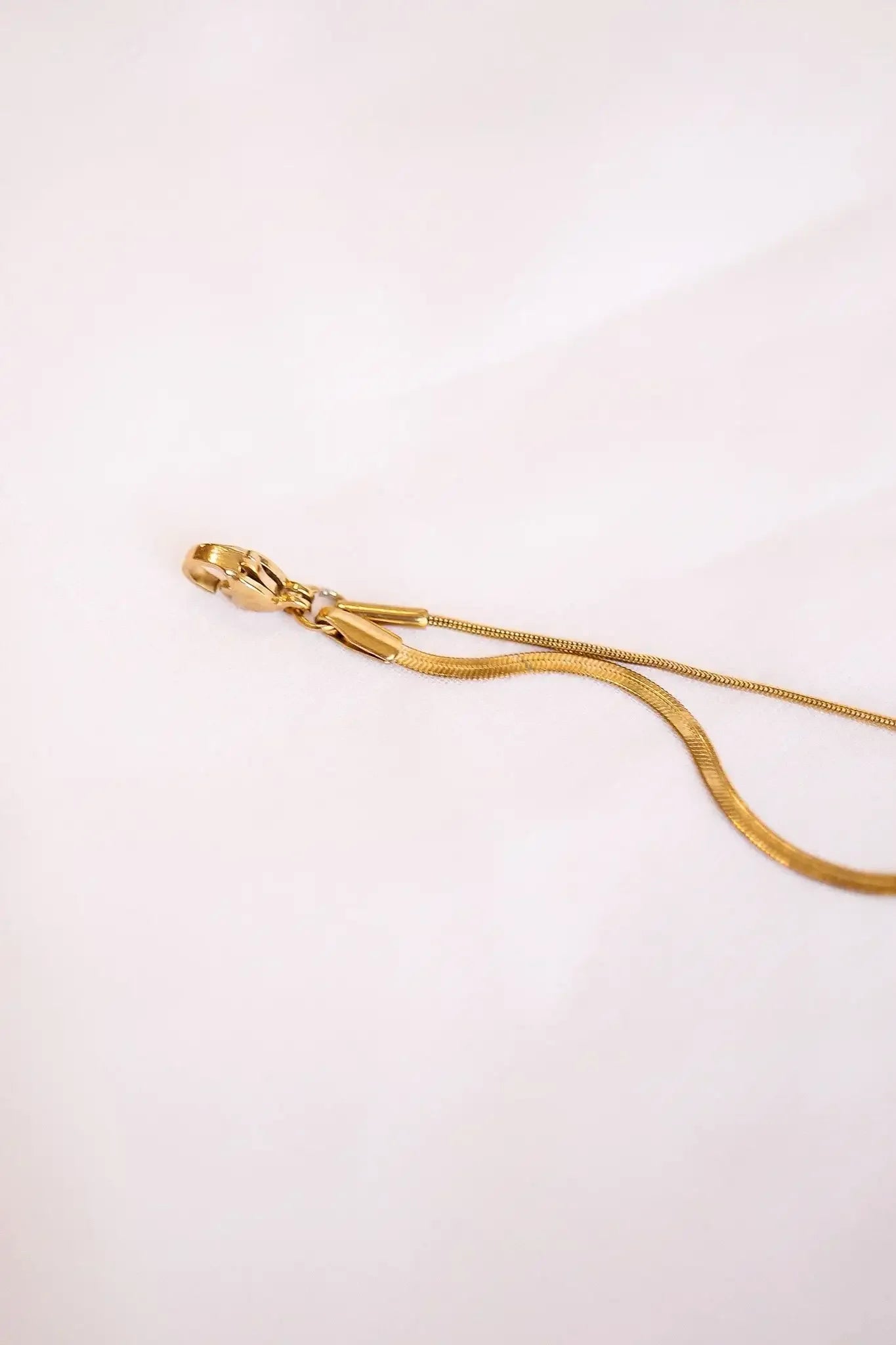 A gold chain with a clasp lies gracefully on a soft, white fabric surface, showcasing its smooth, reflective surface against the pristine backdrop.