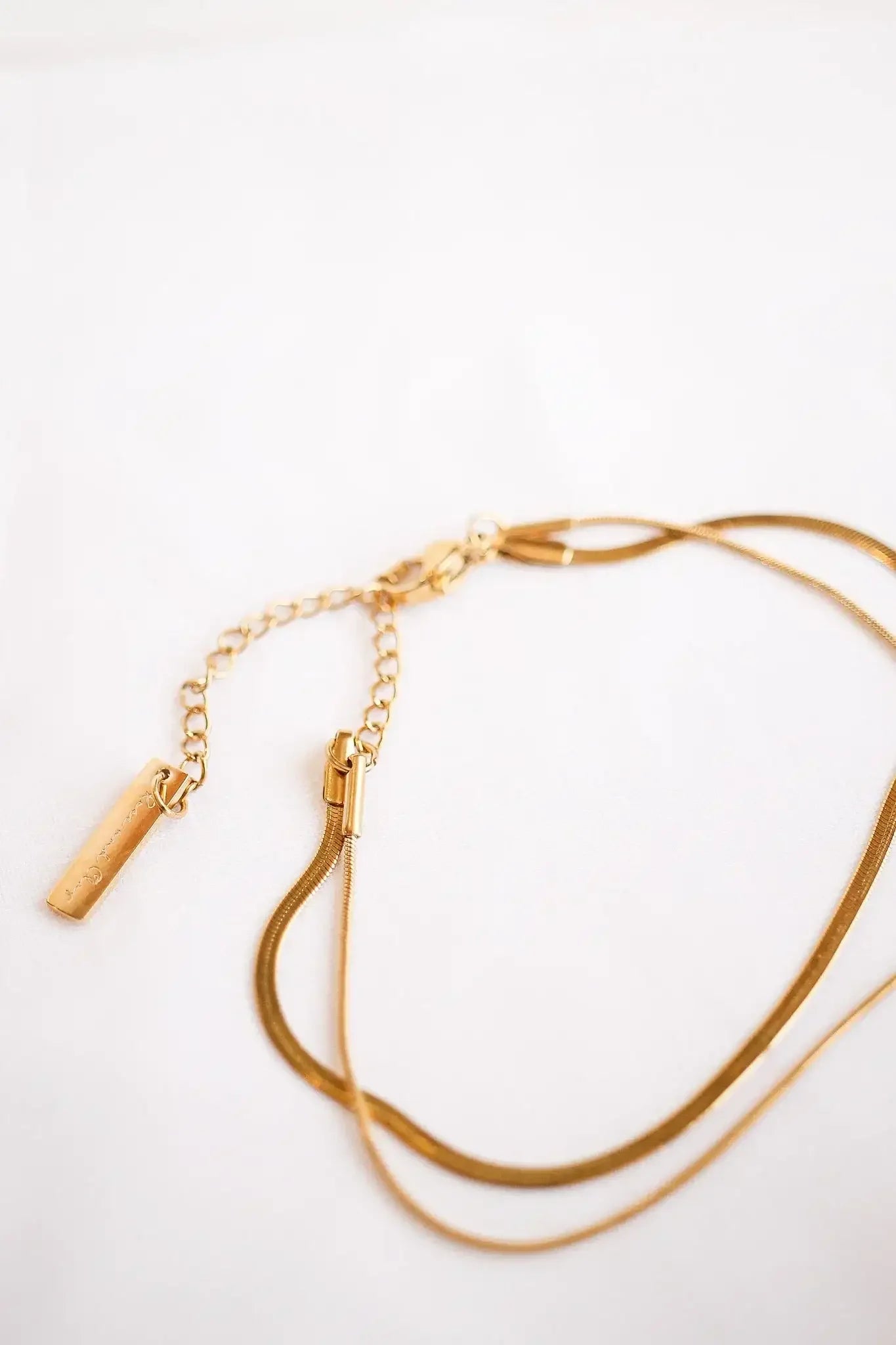 A golden chain necklace rests on a white surface, with a pendant and a clasp visible, showcasing elegant curves and a shiny finish. The overall setting is minimalistic and bright.