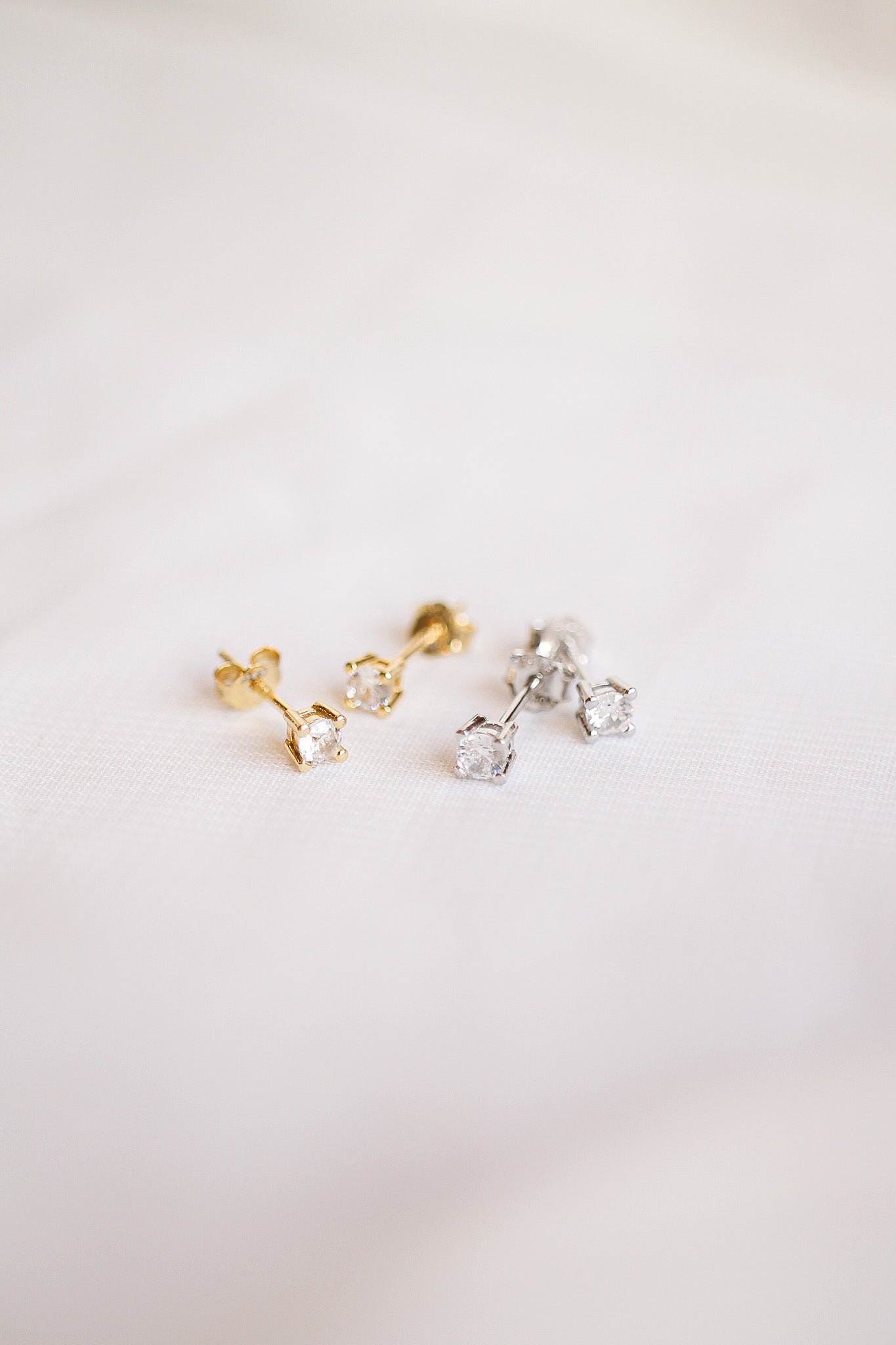 Gold and silver stud earrings, featuring small sparkling gems, rest on a light-colored textured fabric, showcasing their elegance and intricate settings in a soft, diffused light.