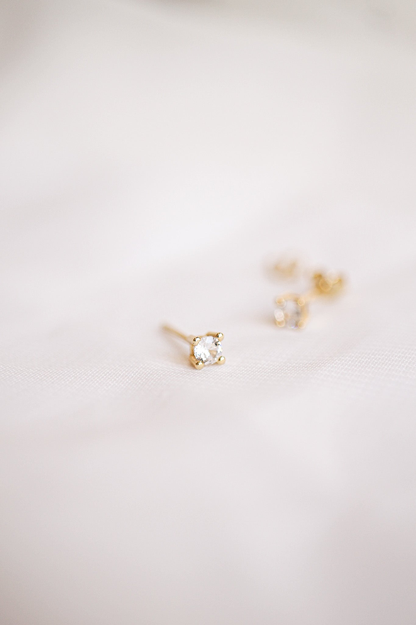Gold stud earrings with small, clear gemstones rest on a soft, light-colored fabric surface, illuminated by gentle lighting that highlights their sparkle and elegance.