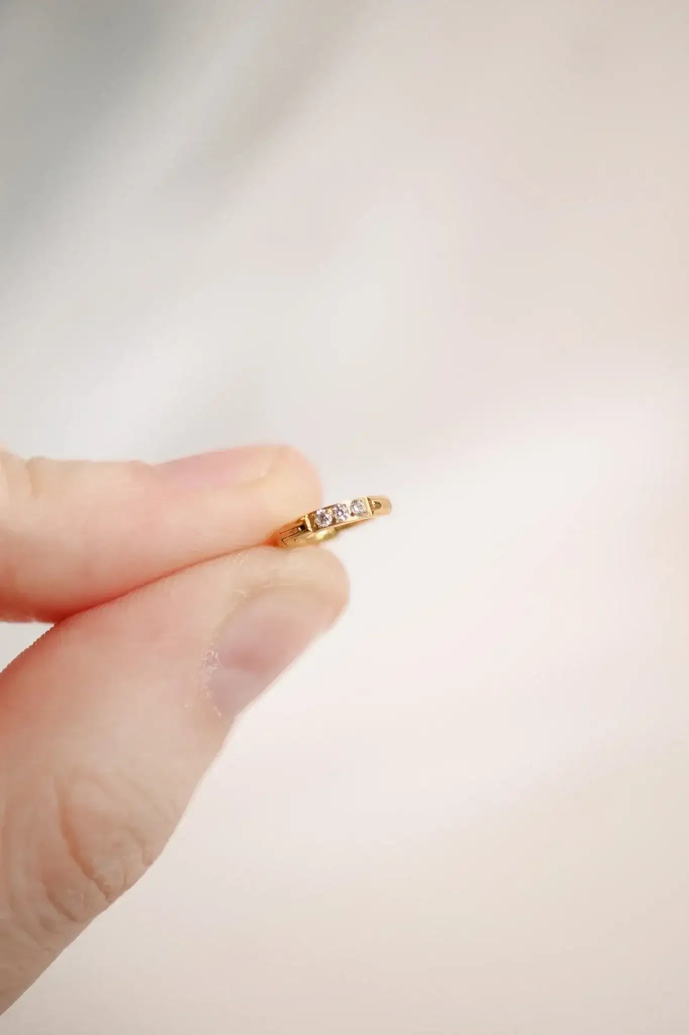 A hand holds a delicate gold ring adorned with three small gemstones. The background is softly blurred, suggesting a calm and neutral setting.