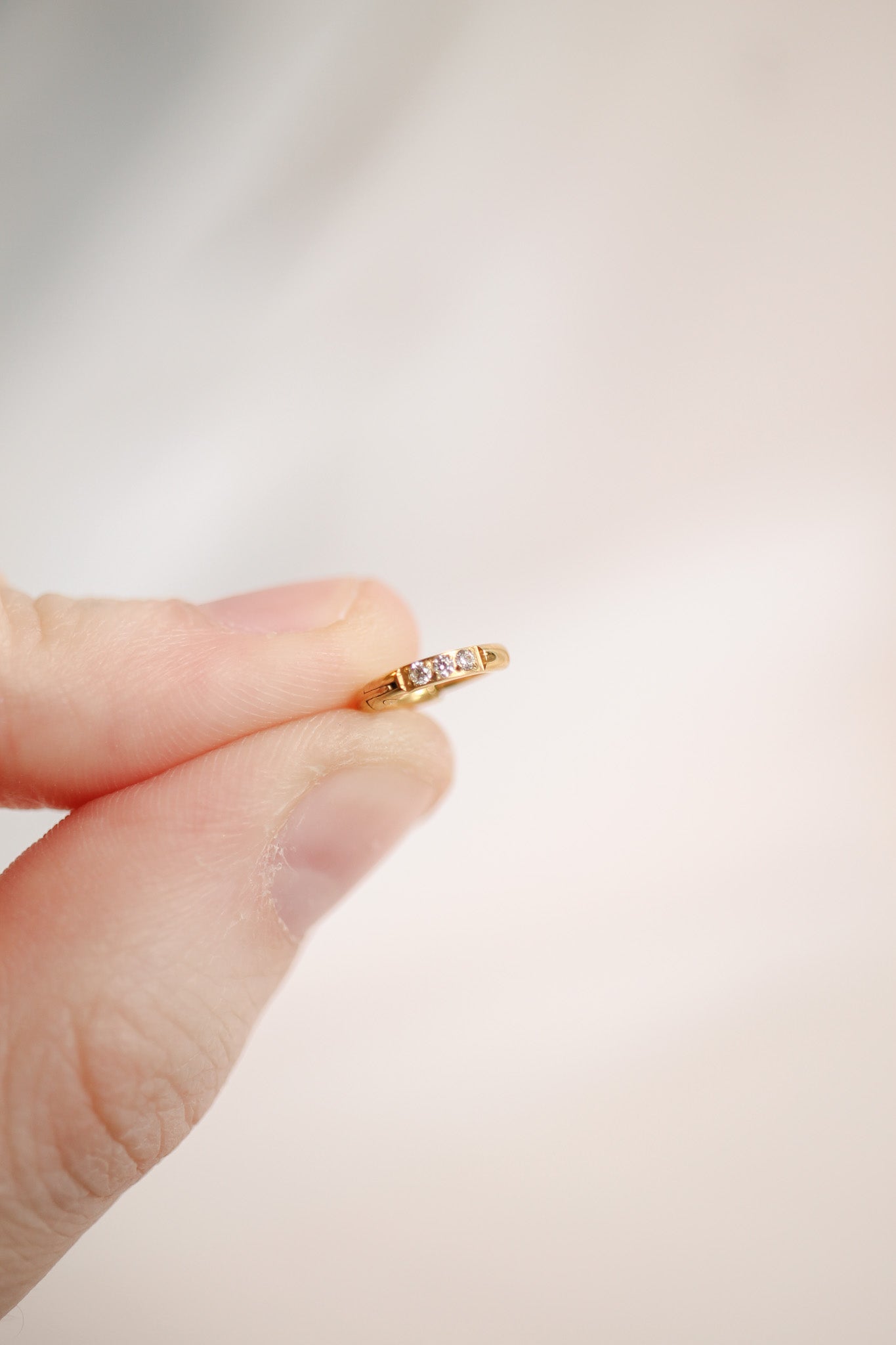 A hand holds a delicate gold ring adorned with three small gemstones. The background is softly blurred, suggesting a calm and neutral setting.