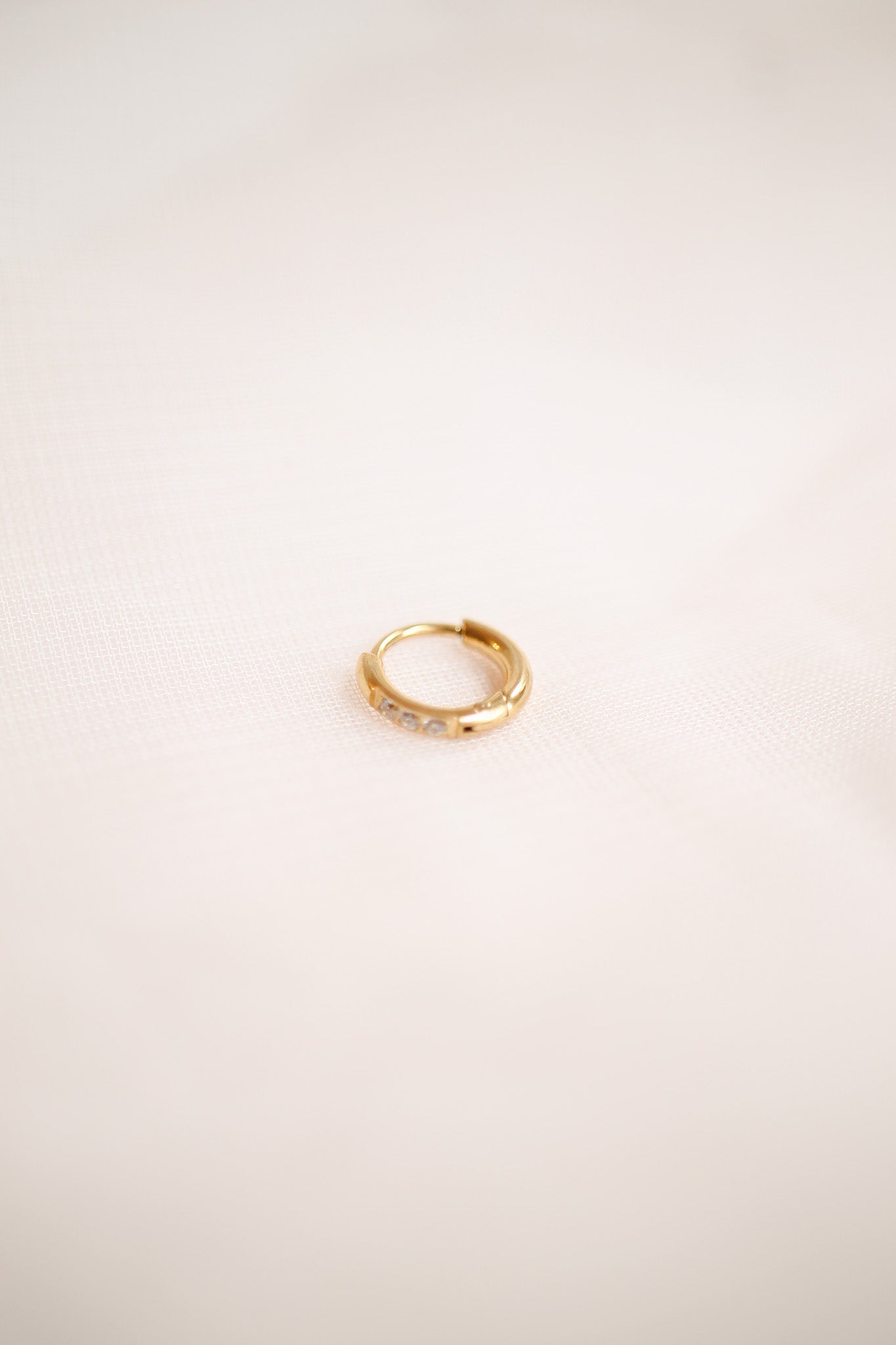 A small gold hoop earring with a smooth surface and three clear stones rests on a soft, light-colored fabric background, highlighting its elegant design and shine.