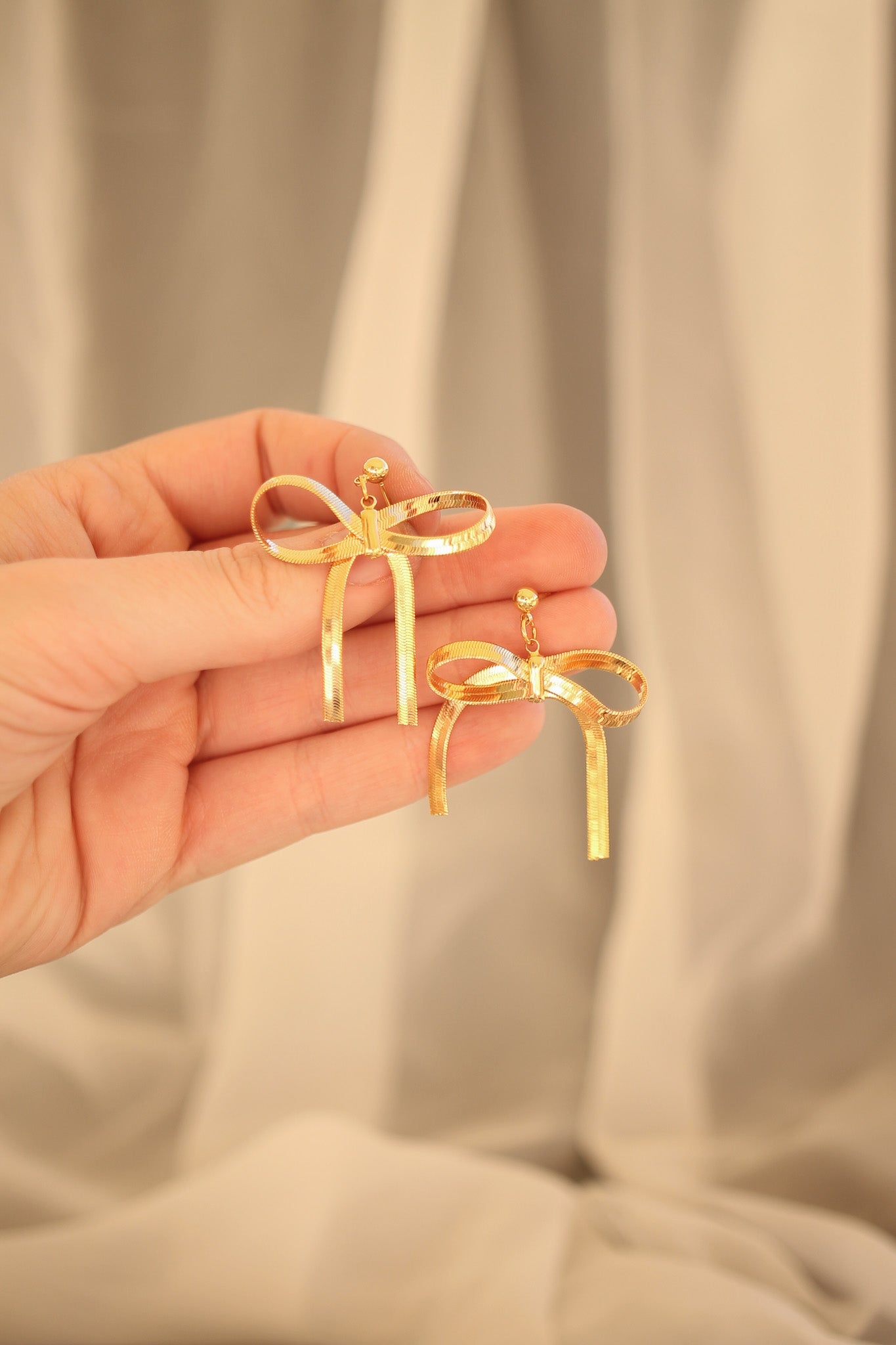 A hand holds two pairs of gold bow-shaped earrings, showcasing their decorative ribbons and clasps against a softly draped, neutral-colored background.