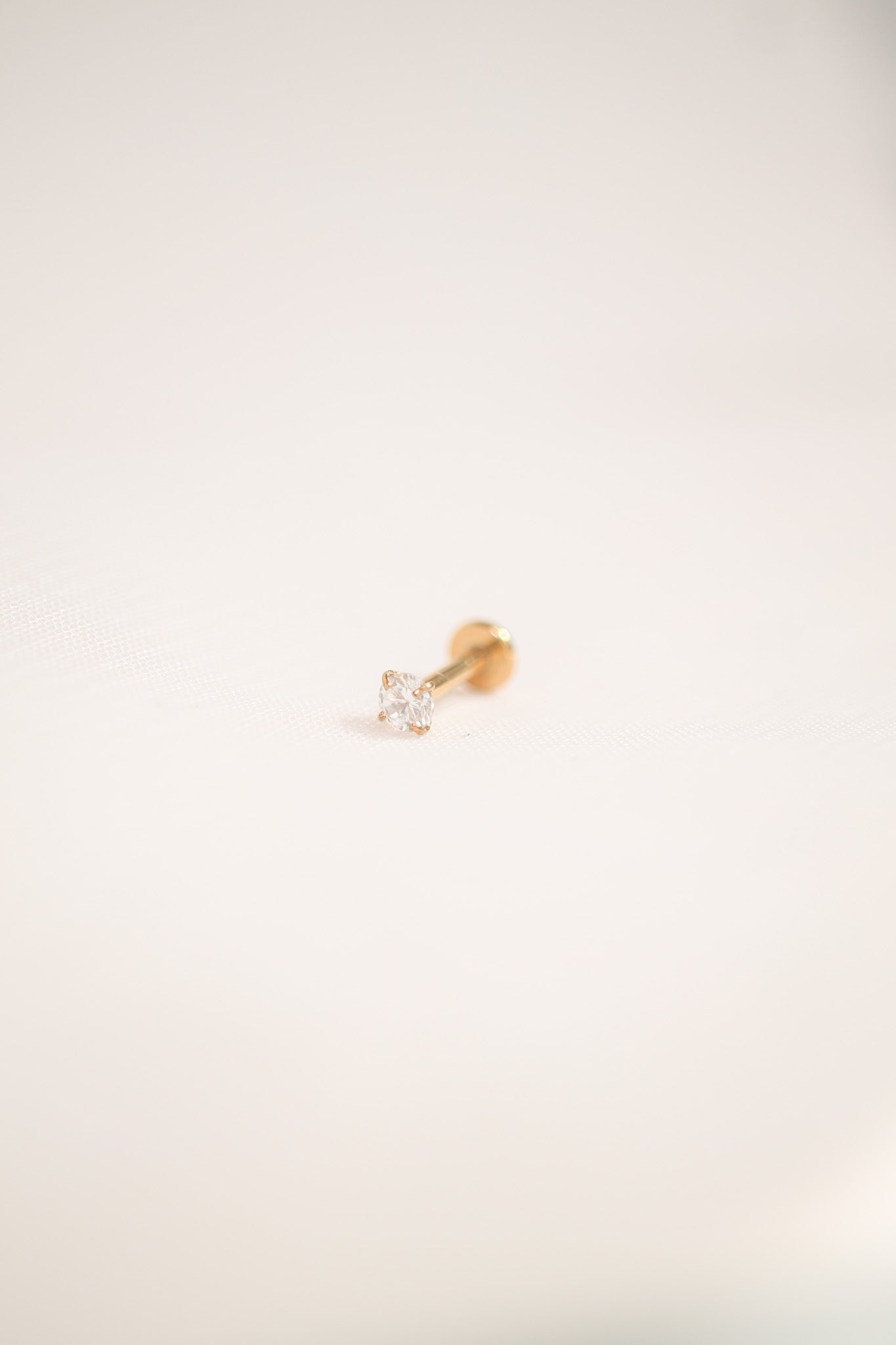 A small, golden stud earring with a sparkling clear gemstone sits flat on a soft, off-white surface, highlighting its delicate design against the neutral backdrop.