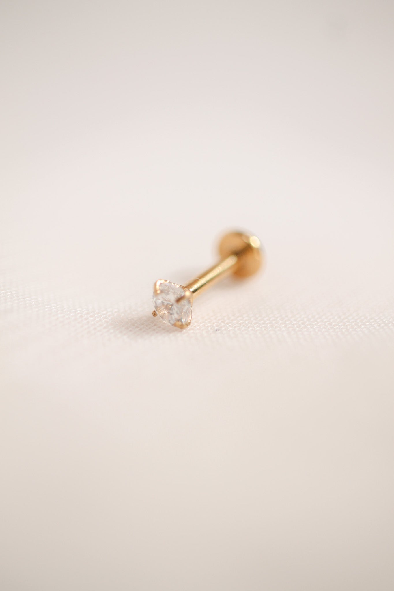 A gold-studded earring lies on a soft, light fabric surface, showcasing a clear, diamond-shaped gemstone at its end, reflecting light subtly in a minimalistic setting.