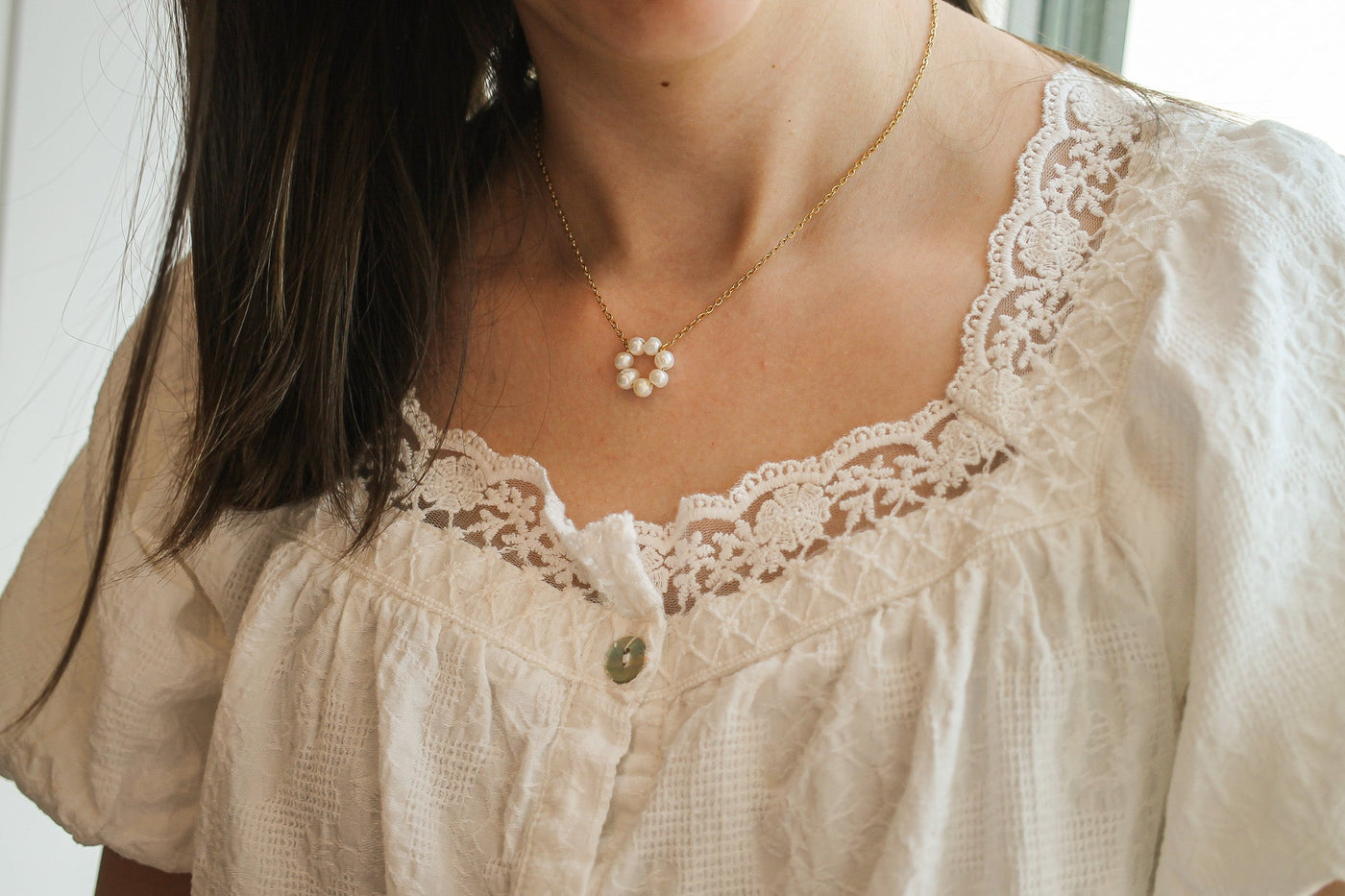 A woman is wearing a delicate gold necklace with a floral pattern made of small pearls. She is dressed in a light, textured white top with lace detailing around the neckline.