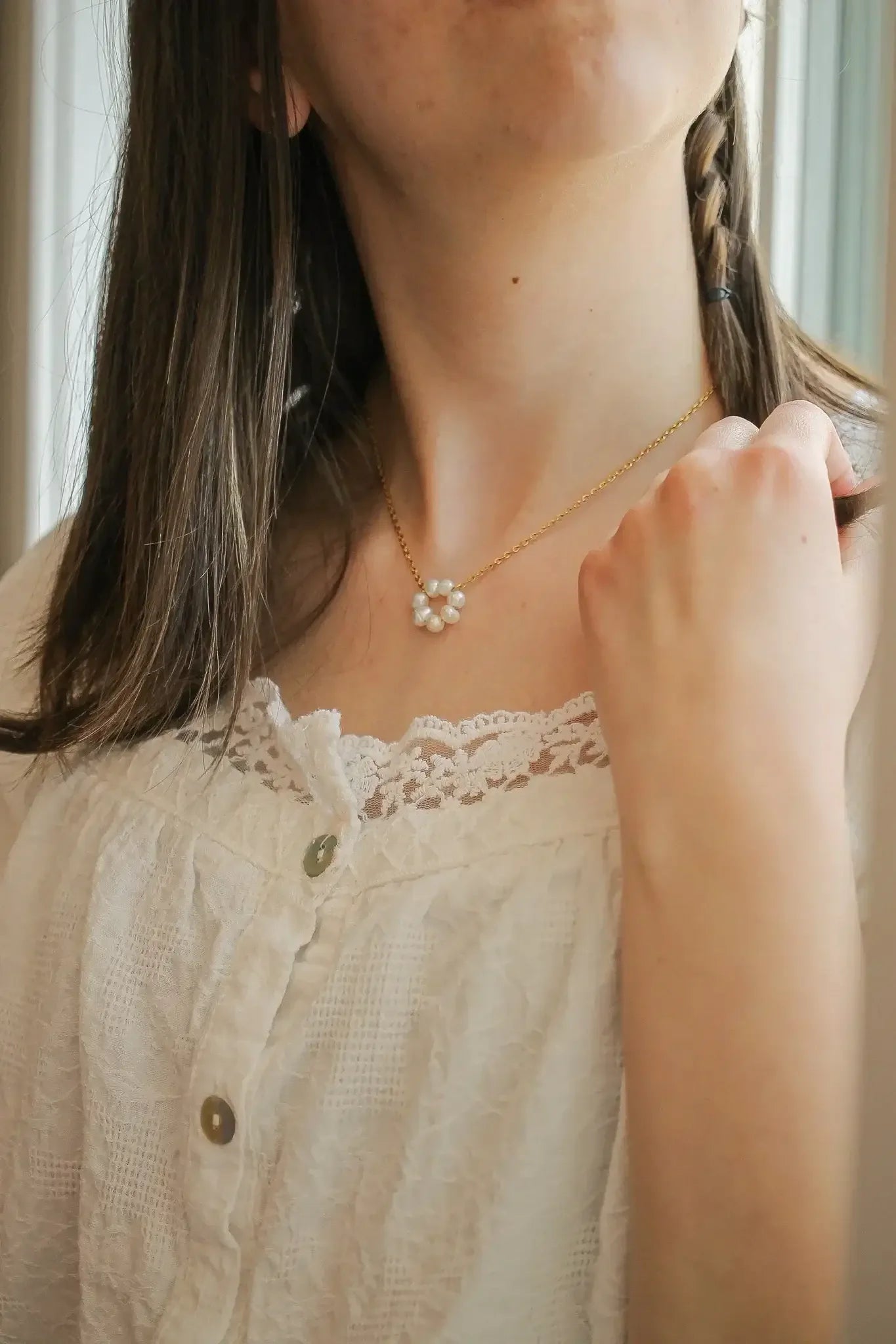 A young woman wears a delicate gold necklace with pearl flowers, gazing upward while gently touching her collar. She is dressed in a white, lace-trimmed blouse, with natural light in the background.