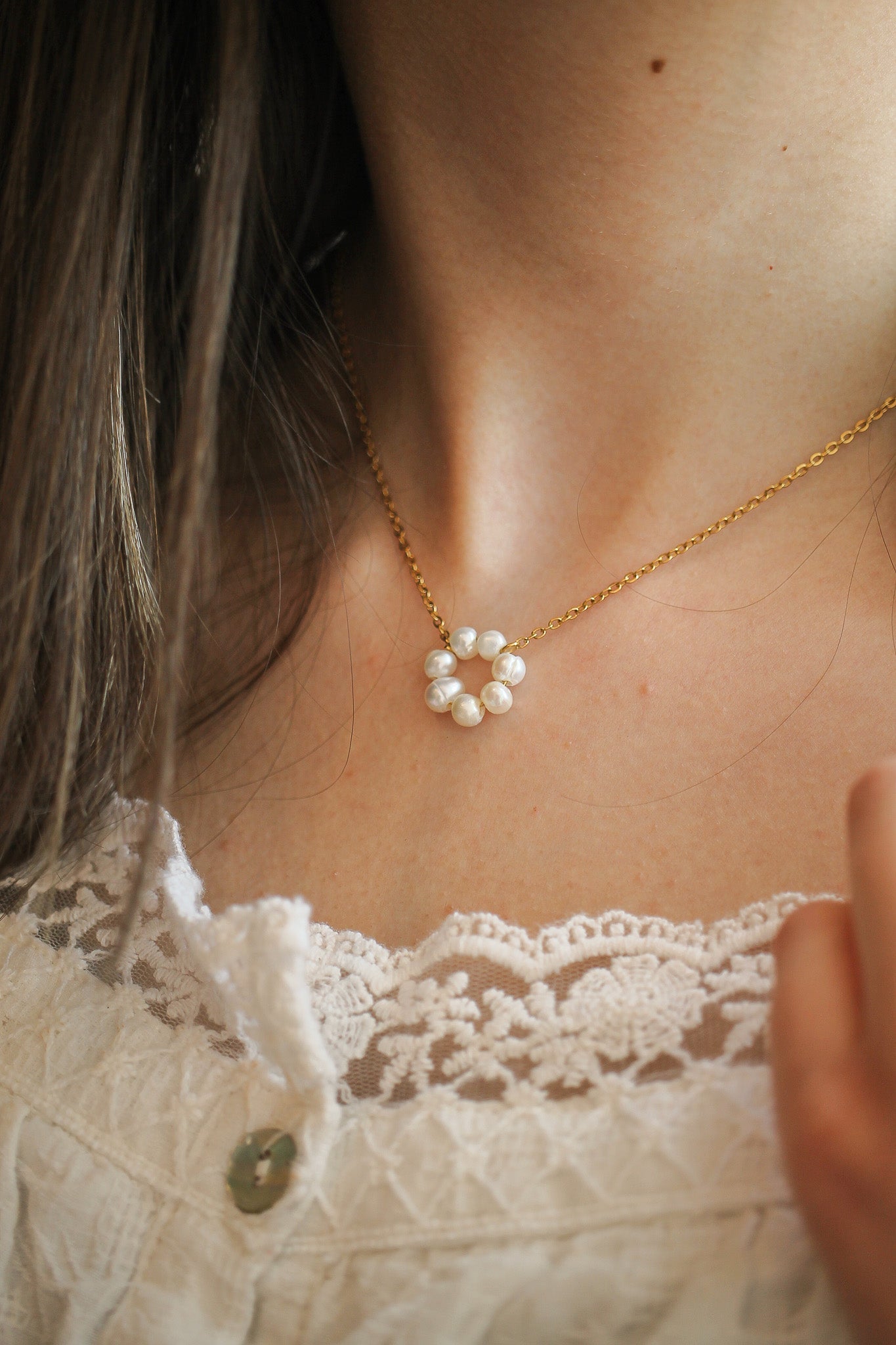 A delicate gold necklace with pearl accents sits around a neck, complementing a white lace top. The wearer gently touches the necklace, highlighting its elegant design.
