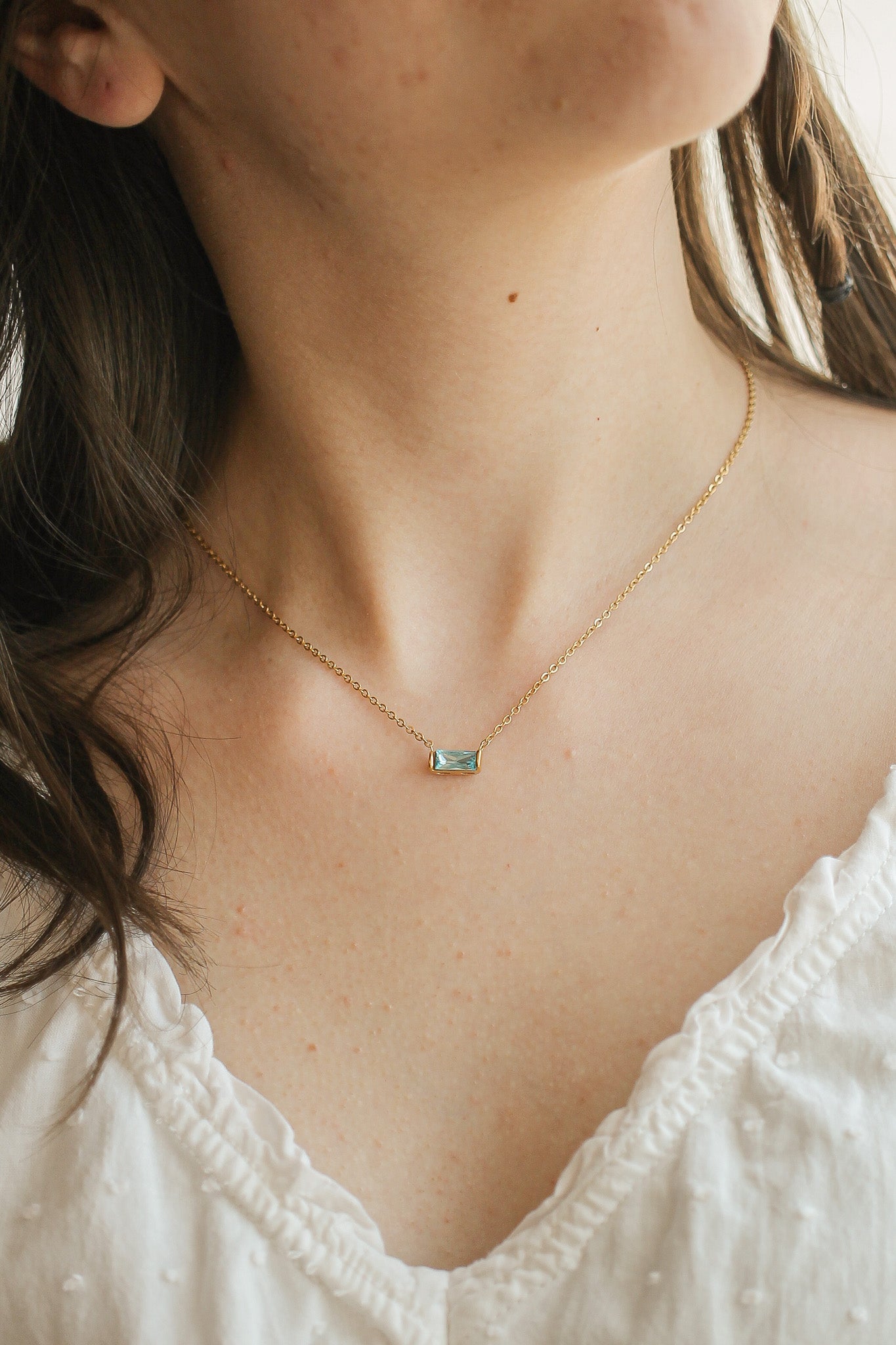A delicate gold necklace with a rectangular gemstone pendant rests on the smooth skin of a person's neck, complementing their simple white blouse. The background is softly blurred.