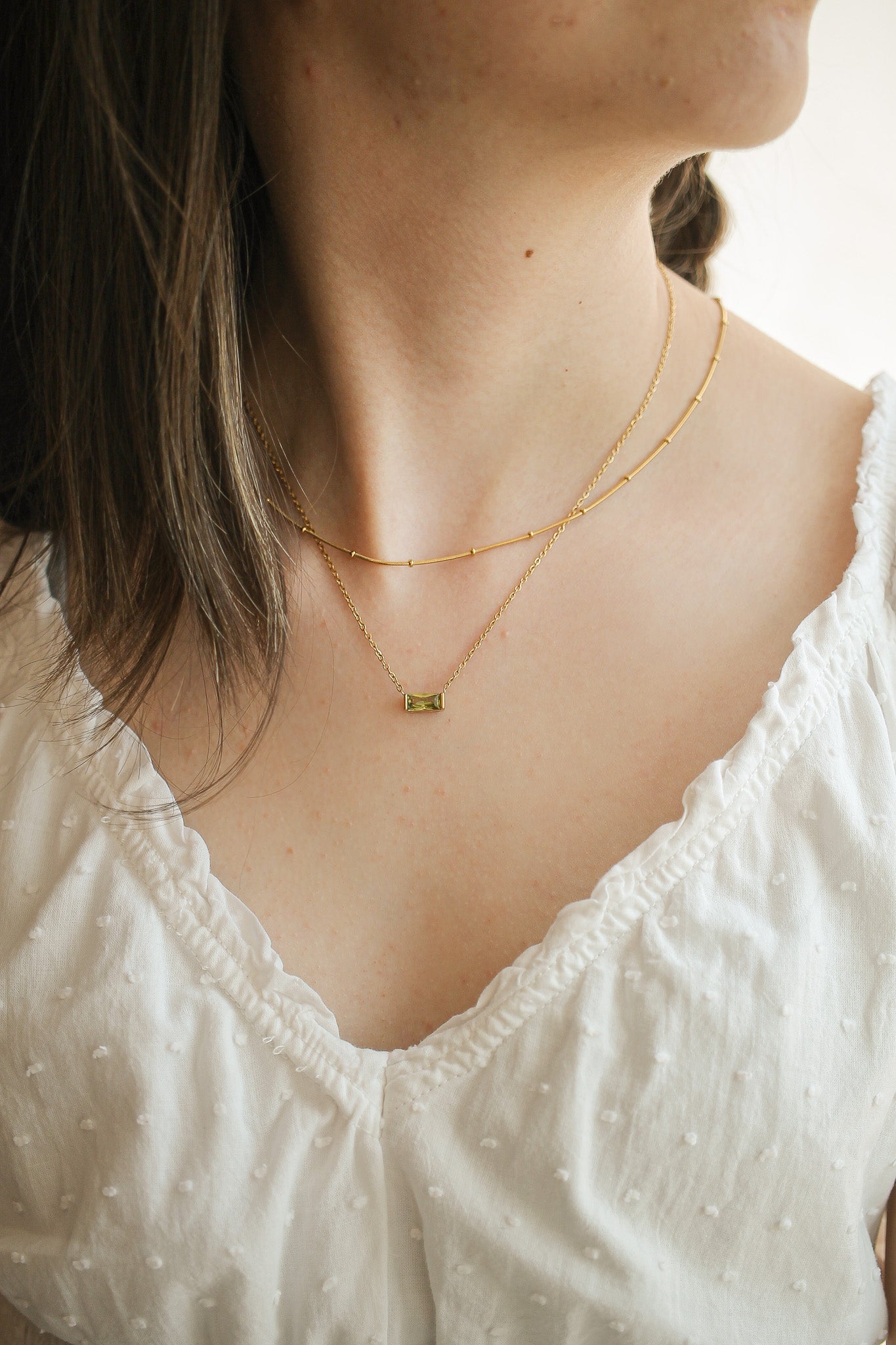 A person wears multiple delicate gold necklaces, including a rectangular pendant, against a light background. The attire features a soft, textured white fabric with small dots.