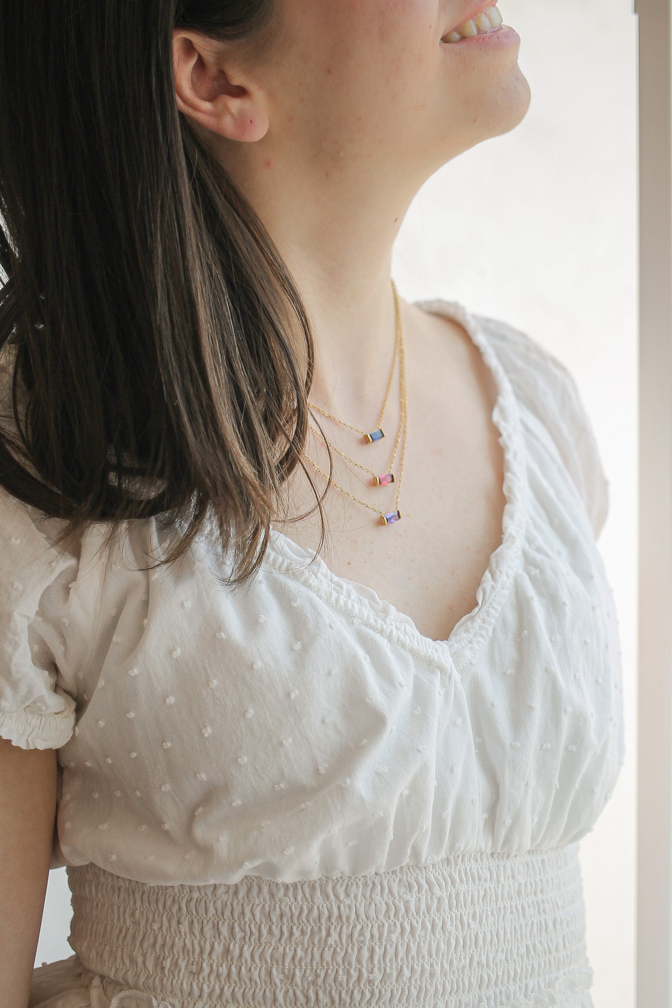 A woman smiles, wearing a white, textured top adorned with a soft neckline. She displays layered necklaces with colorful accents, standing in a softly lit indoor space.