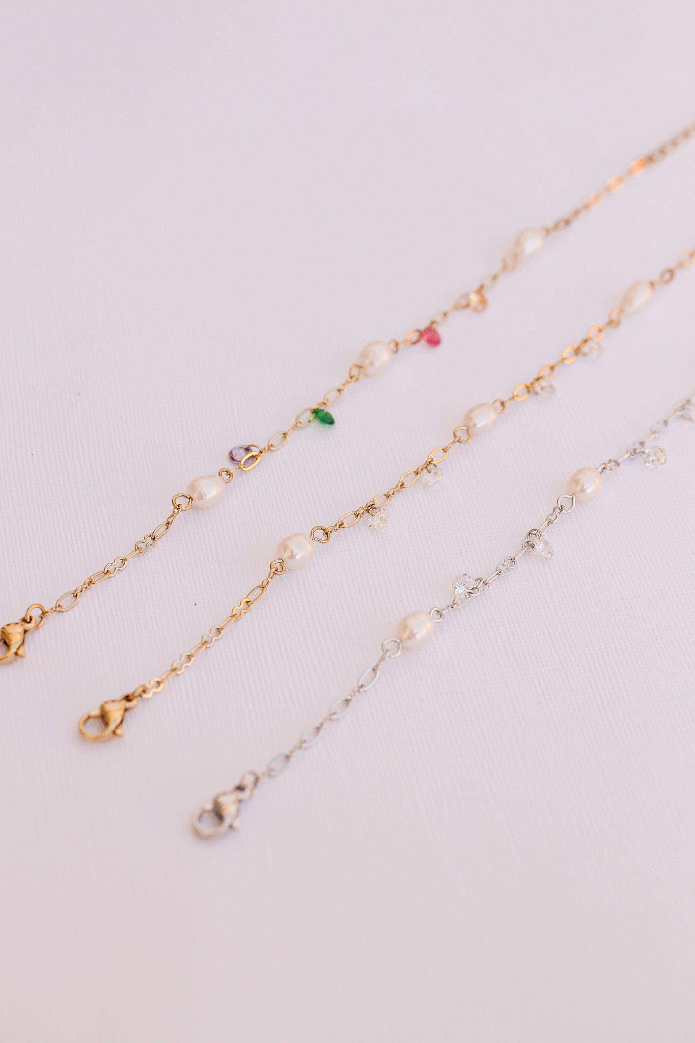 Three delicate chains composed of gold and silver are adorned with pearls and colorful gemstones. They lie flat on a light-colored surface, showcasing their elegant designs and clasps.