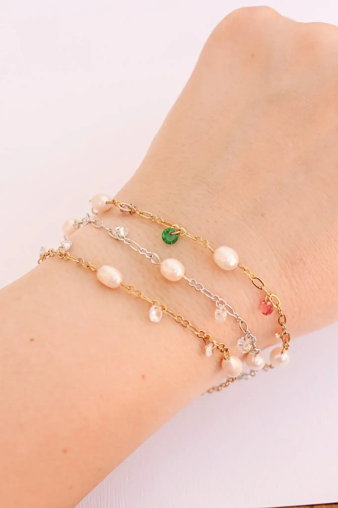 A wrist adorned with three delicate bracelets: one golden, one silver, and one mixed with pearls and colorful gems, resting against a light, textured background.