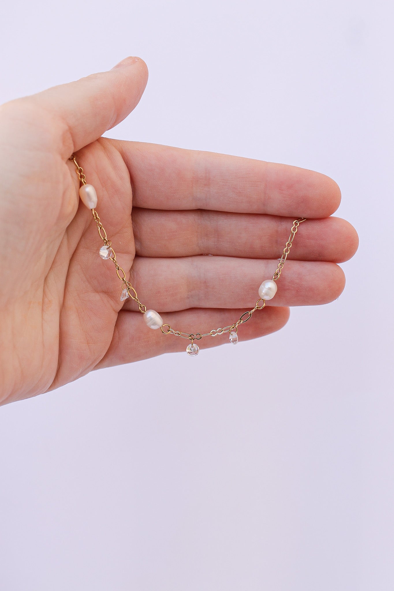 A hand holds a delicate gold chain necklace adorned with pearls and crystal beads against a plain, light background, showcasing its intricate design and elegance.