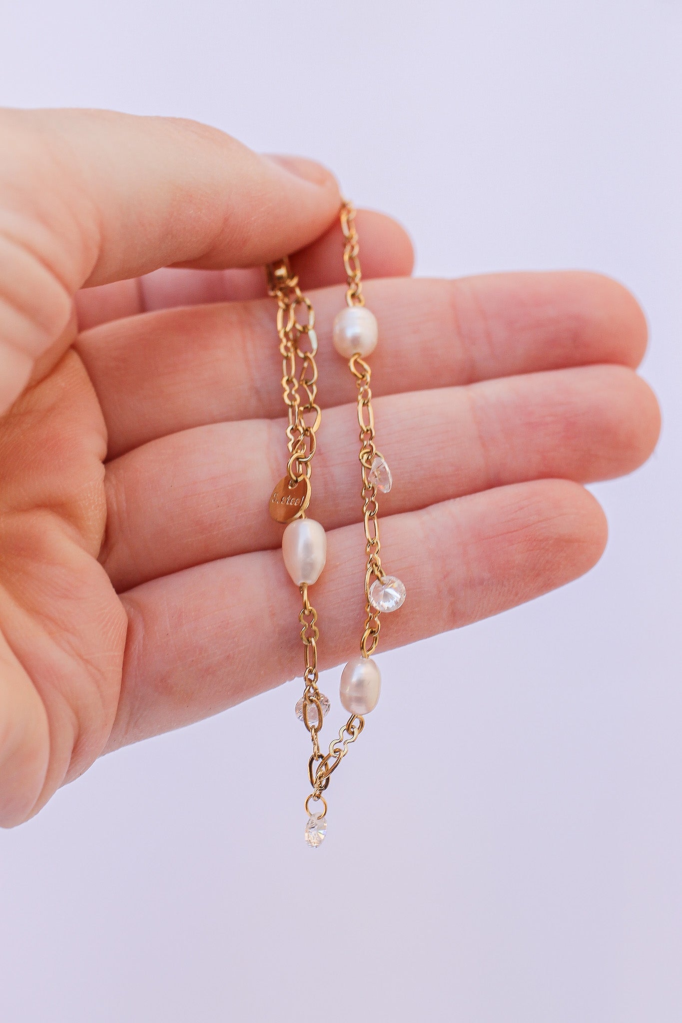 A hand holds a delicate gold chain necklace featuring several pearls and small crystals, displayed against a light background, suggesting elegance and careful craftsmanship.