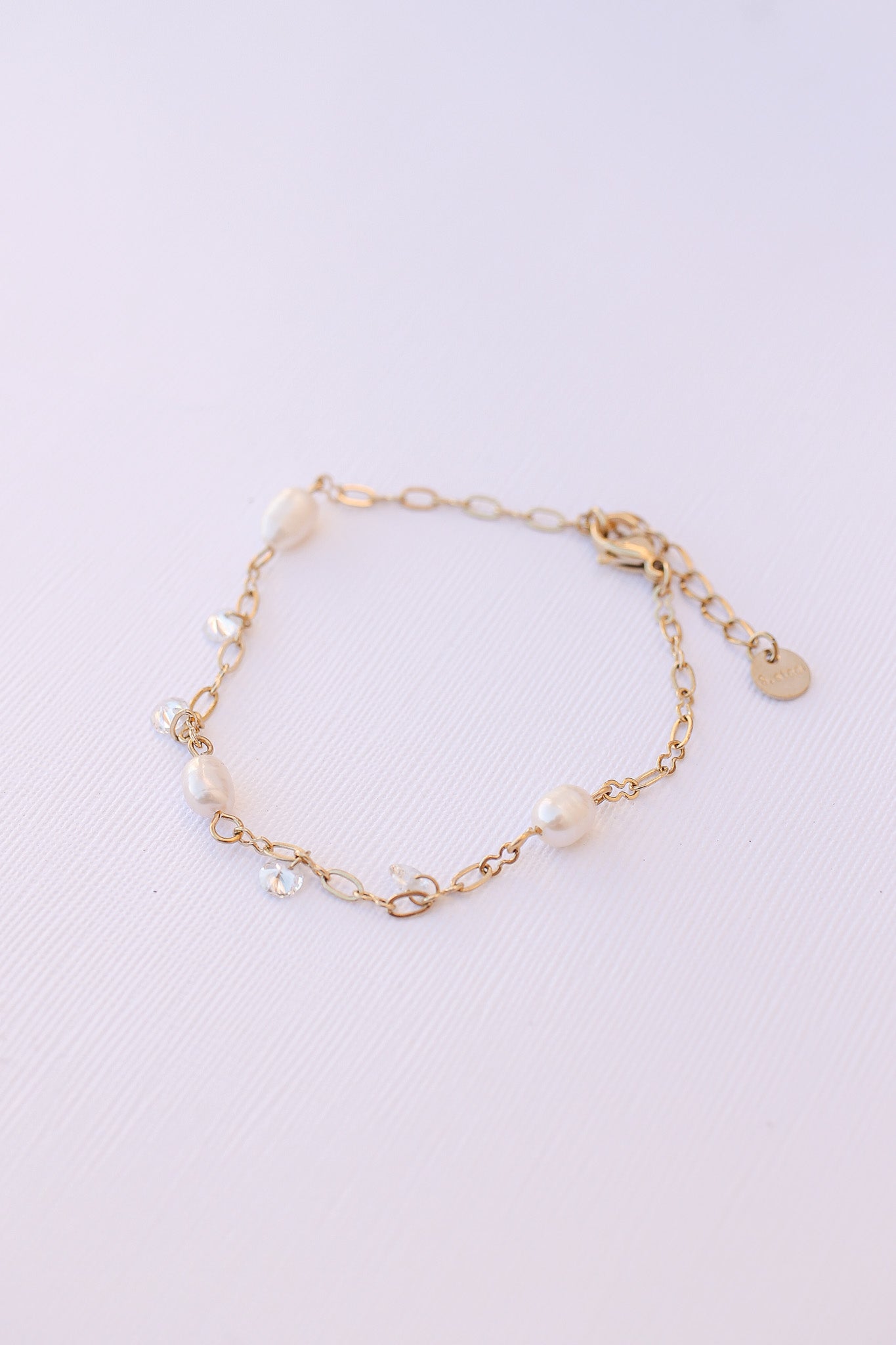 A delicate gold bracelet features lustrous pearls and small crystals linked together. It rests flat on a textured white surface, showcasing its elegant design and materials.