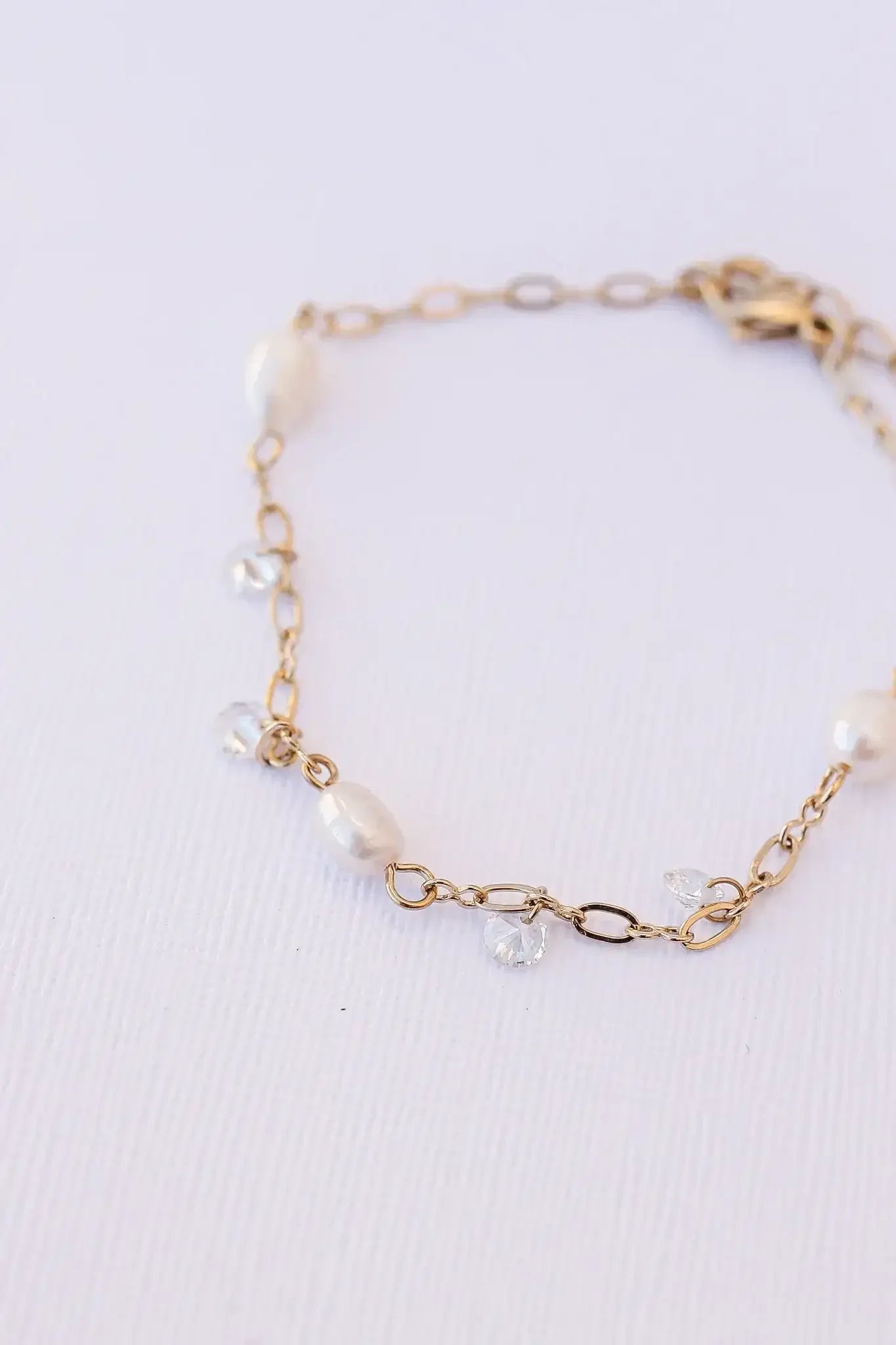 A delicate gold bracelet featuring smooth white pearls and sparkling clear crystals, resting on a textured white surface, showcasing its elegant design and craftsmanship.