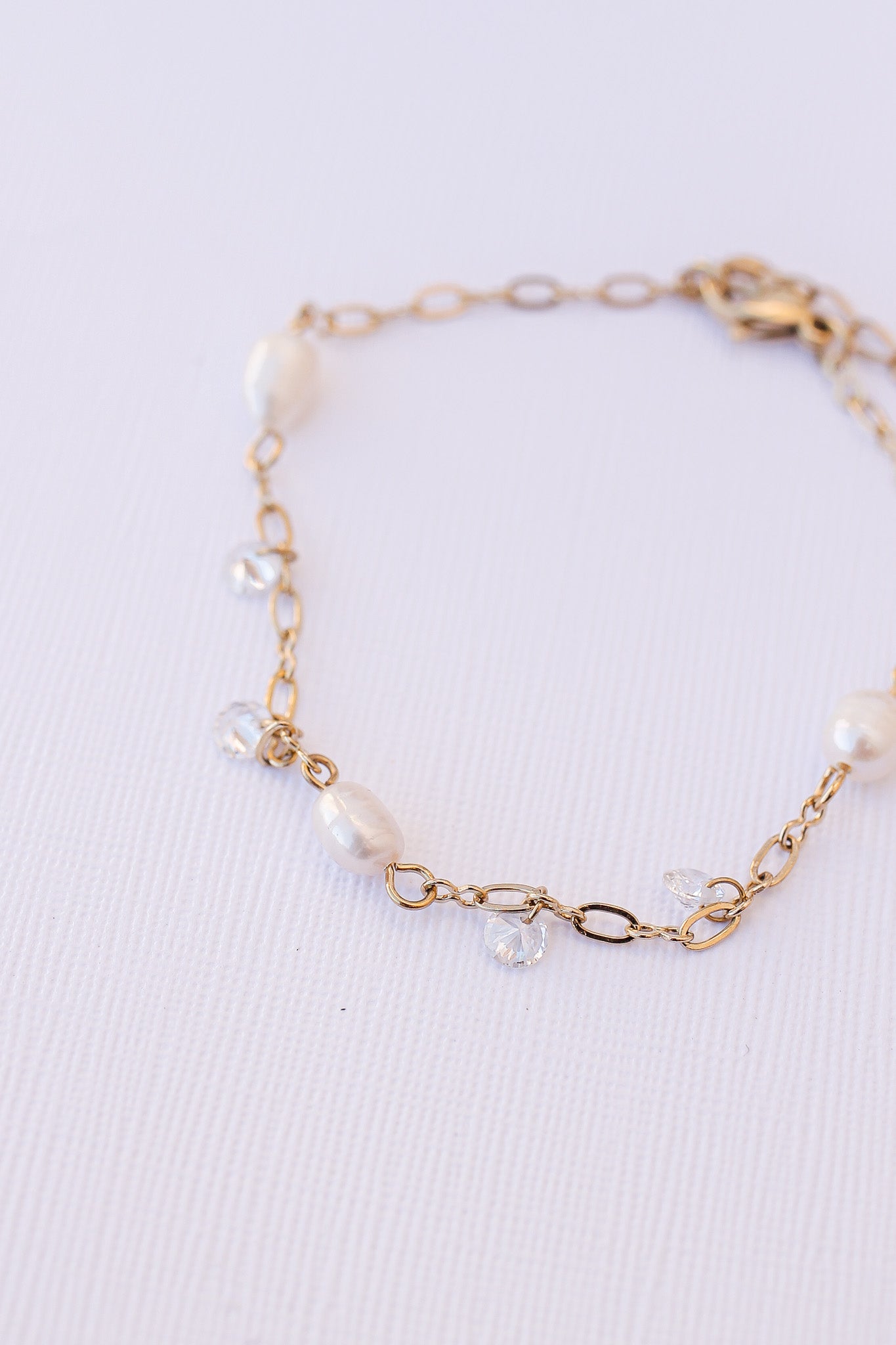 A delicate gold bracelet featuring smooth white pearls and sparkling clear crystals, resting on a textured white surface, showcasing its elegant design and craftsmanship.