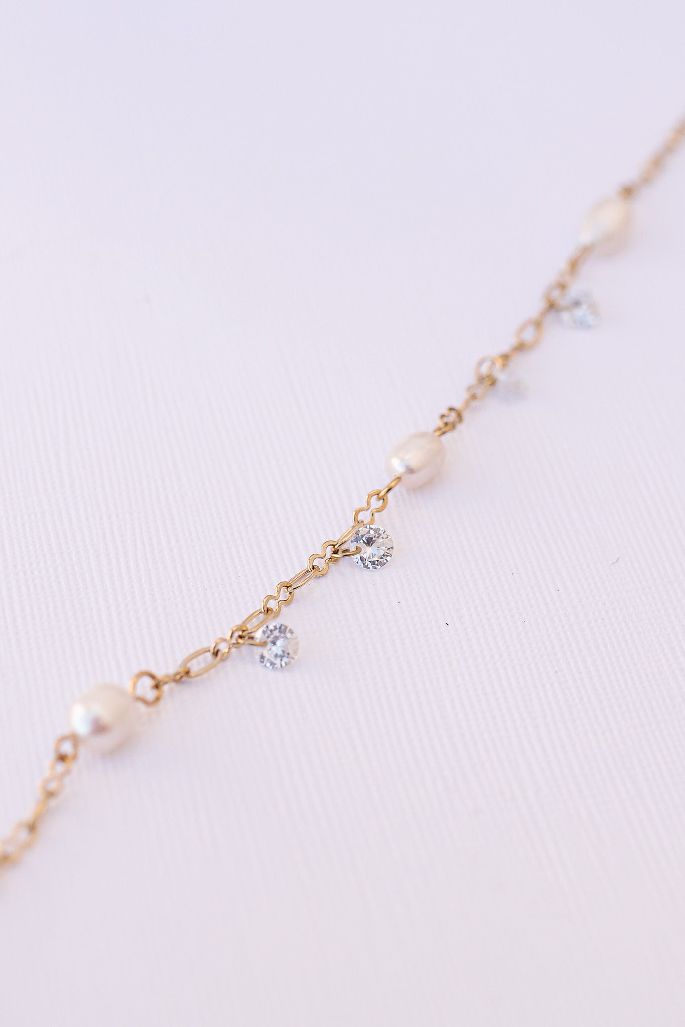 A delicate gold chain features alternating pearls and sparkling rhinestones, arranged symmetrically. The chain rests gently against a soft, textured white background, highlighting its elegance.