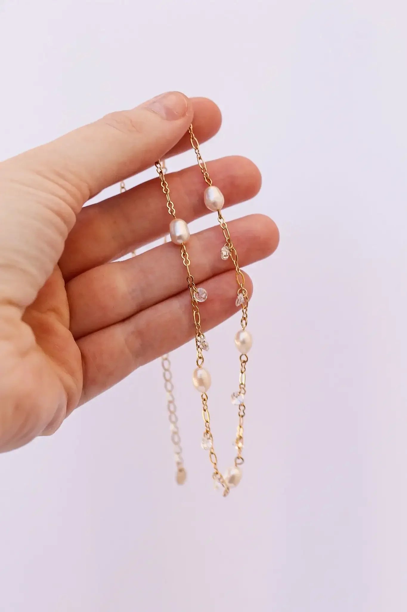 A hand holds a delicate gold chain adorned with pearls and small crystals. The background is light and neutral, emphasizing the jewelry's elegance and detail.
