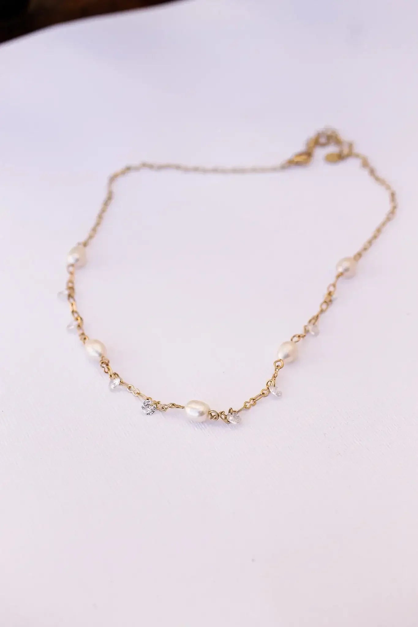 A delicate gold necklace adorned with pearls and small gemstones lies on a textured white surface, showcasing its elegant design and intricate detailing in a softly lit setting.