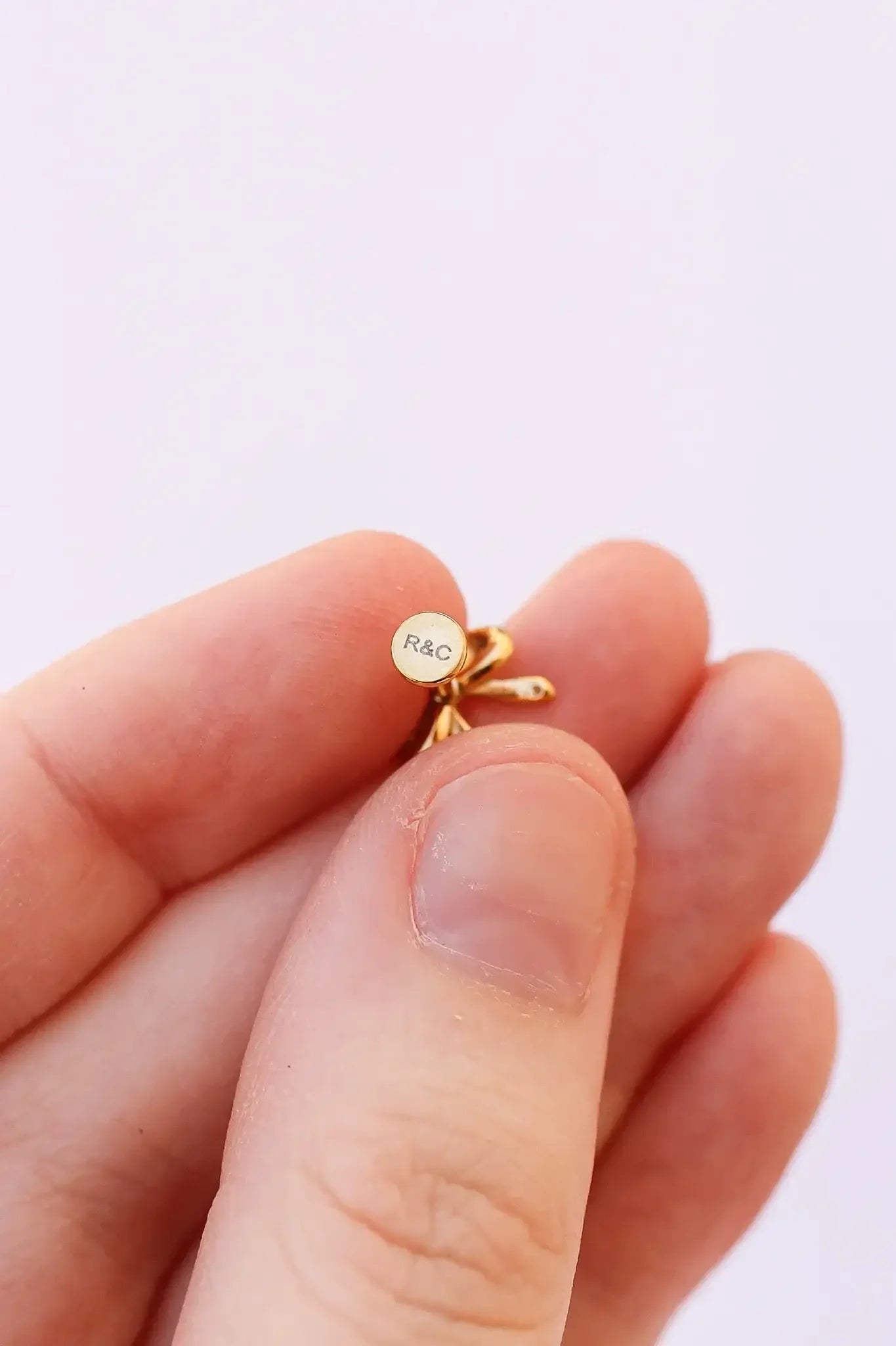 A hand holds a small, golden earring featuring a circular component with the inscription "R&C." The background is plain and light-colored, emphasizing the earring's details.