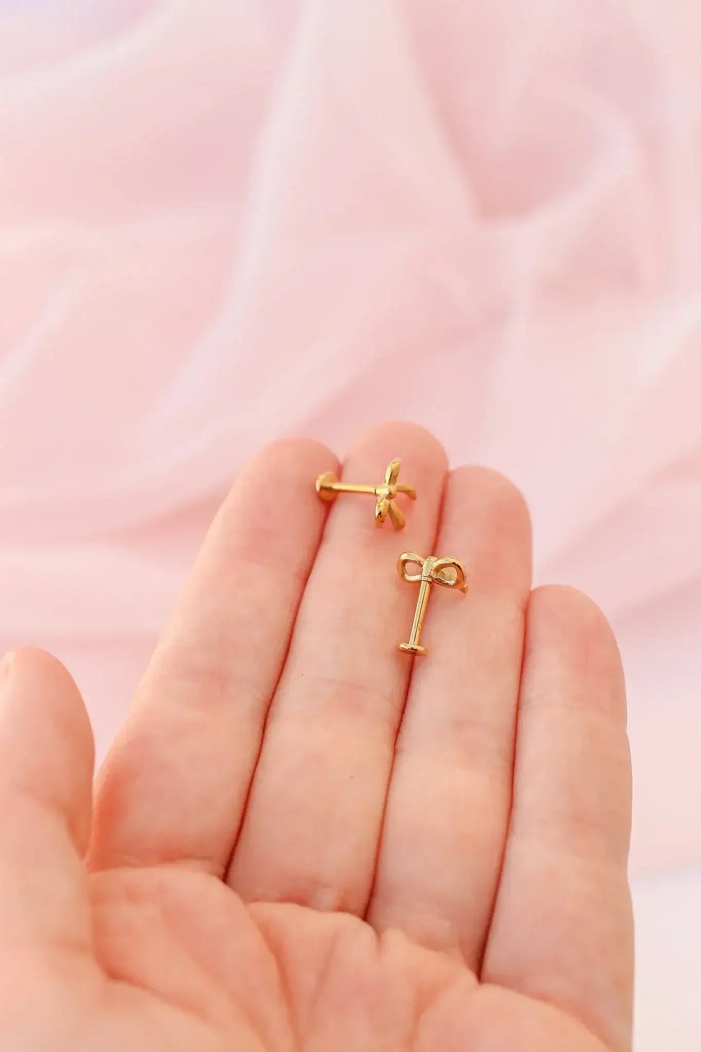 A hand holds two gold-colored earrings shaped like bows and flowers. They rest against a soft, pink, fabric background, suggesting a delicate, elegant setting.