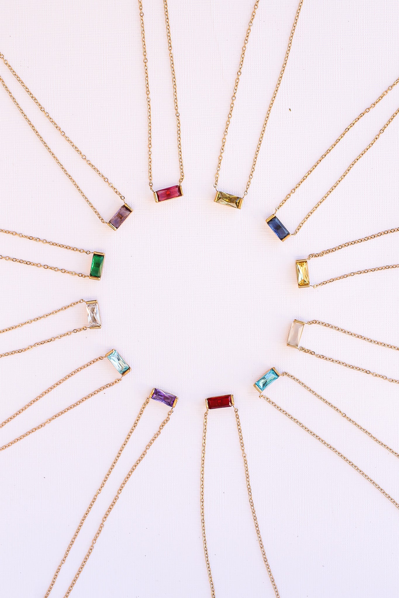 Colorful rectangular gemstone necklaces are arranged in a circular pattern. Each necklace features a gold chain and a different colored stone, displayed on a light-colored background.
