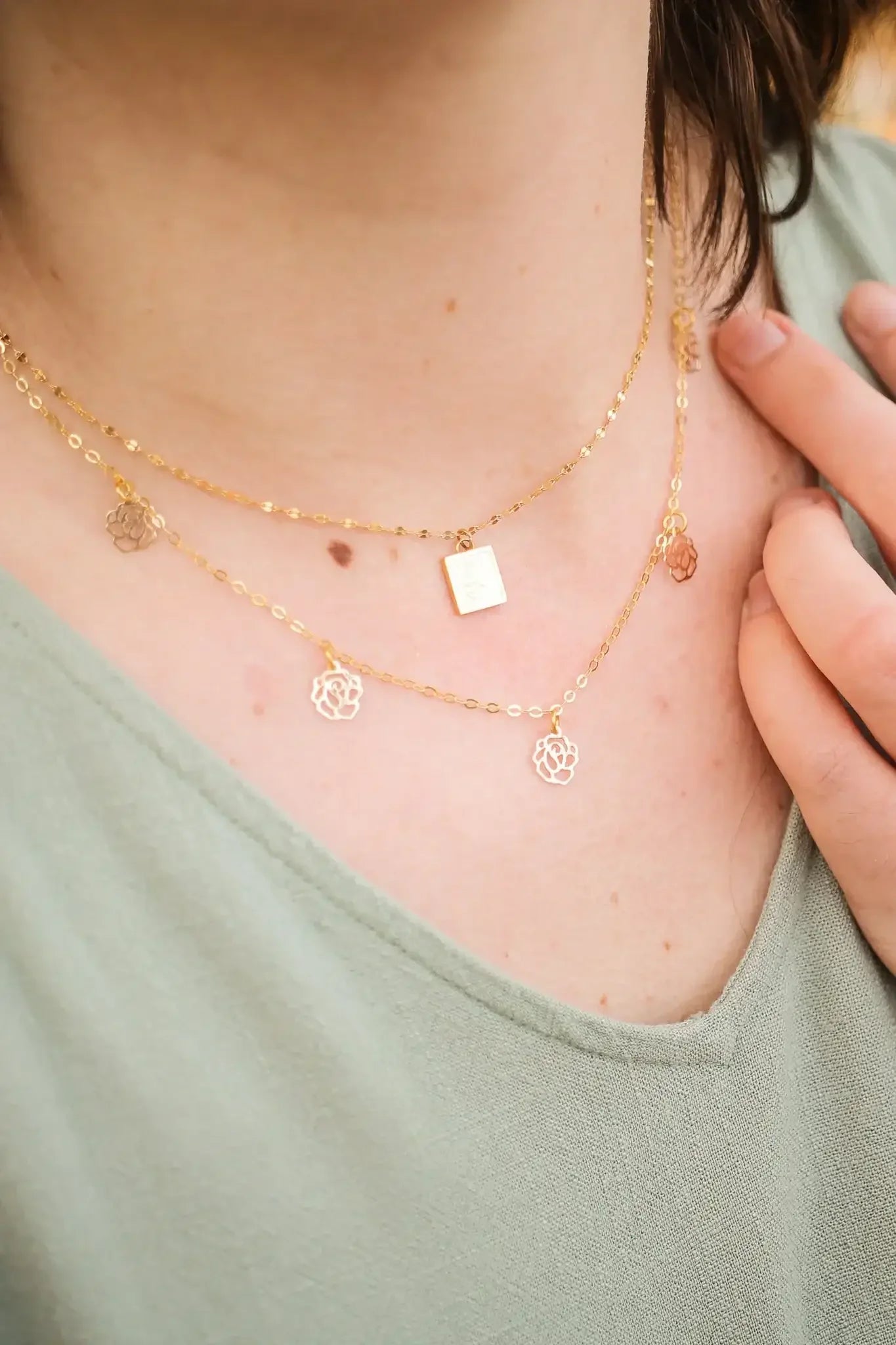 Three delicate gold necklaces adorn a person’s neck, featuring a rectangular charm and flower-shaped pendants. The individual lightly touches one necklace while wearing a green top in a soft, natural setting.