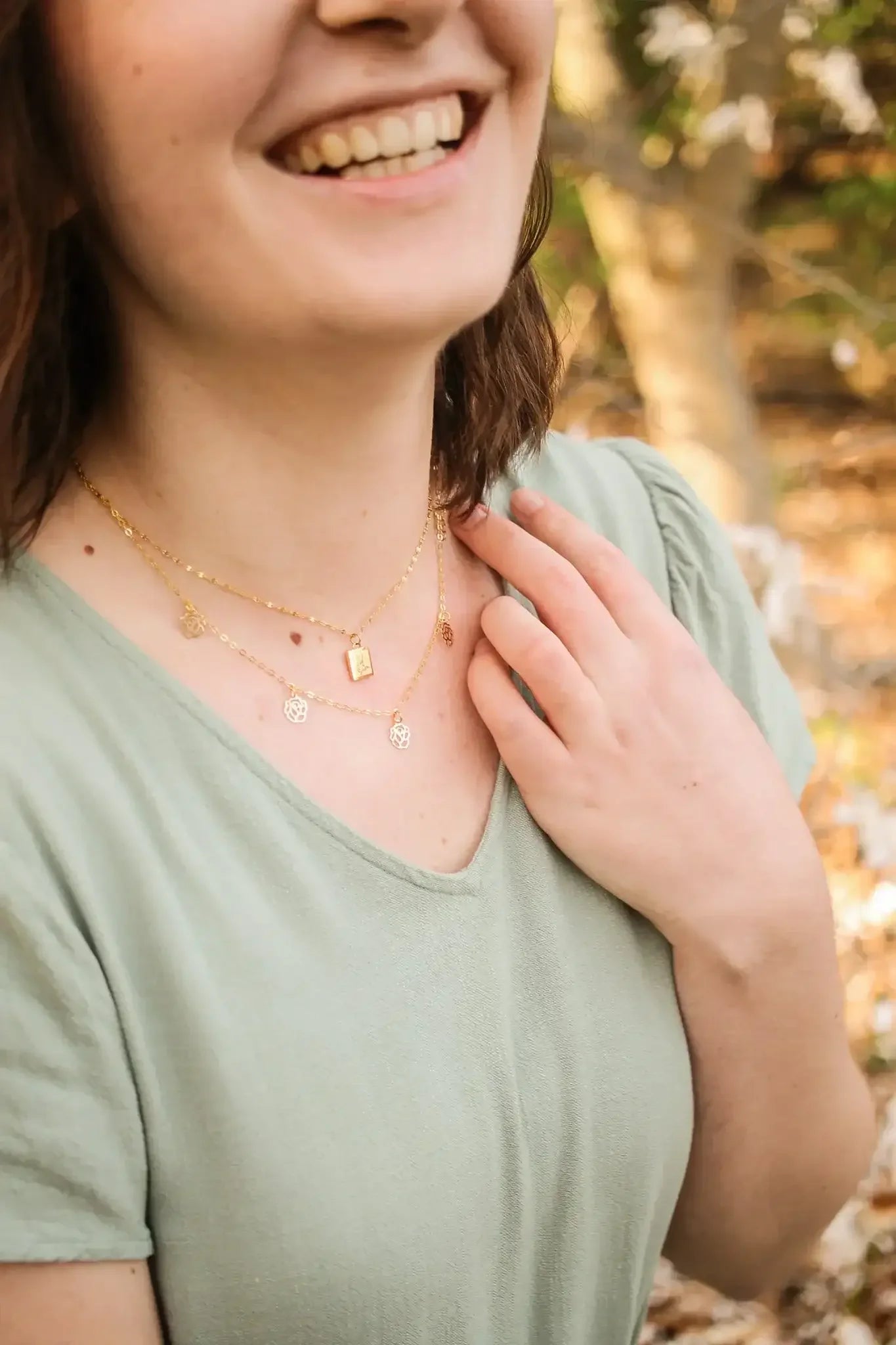 A woman smiles while wearing layered gold necklaces. She gently touches one necklace with her hand in a natural outdoor setting, surrounded by blurred greenery and soft earth tones.