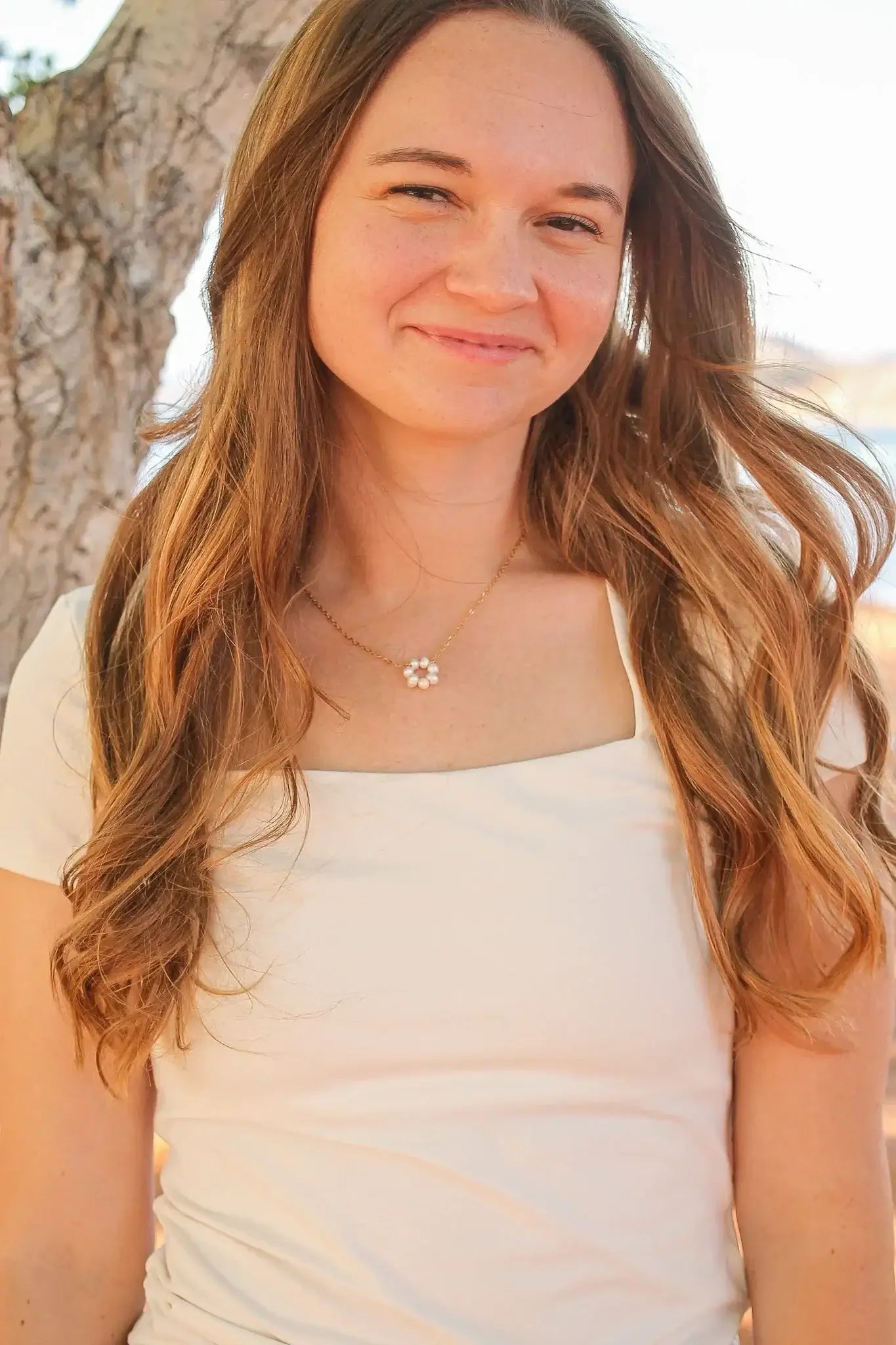 A woman with long, wavy hair smiles warmly while wearing a light-colored shirt and a floral necklace. She stands near a tree with a blurred outdoor background.
