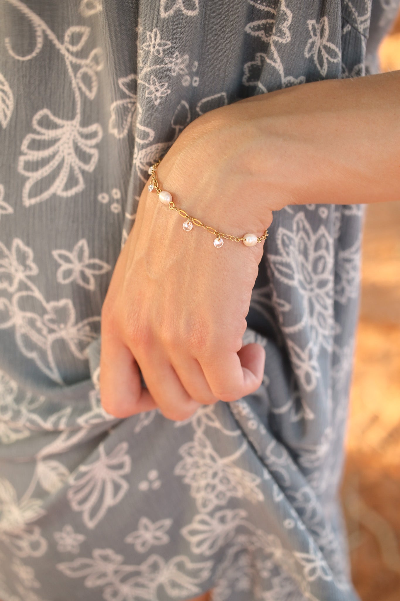 A delicate gold bracelet with pearls and small gems adorns a woman's wrist, while her hand gently holds fabric patterned with white floral designs in a softly lit outdoor setting.