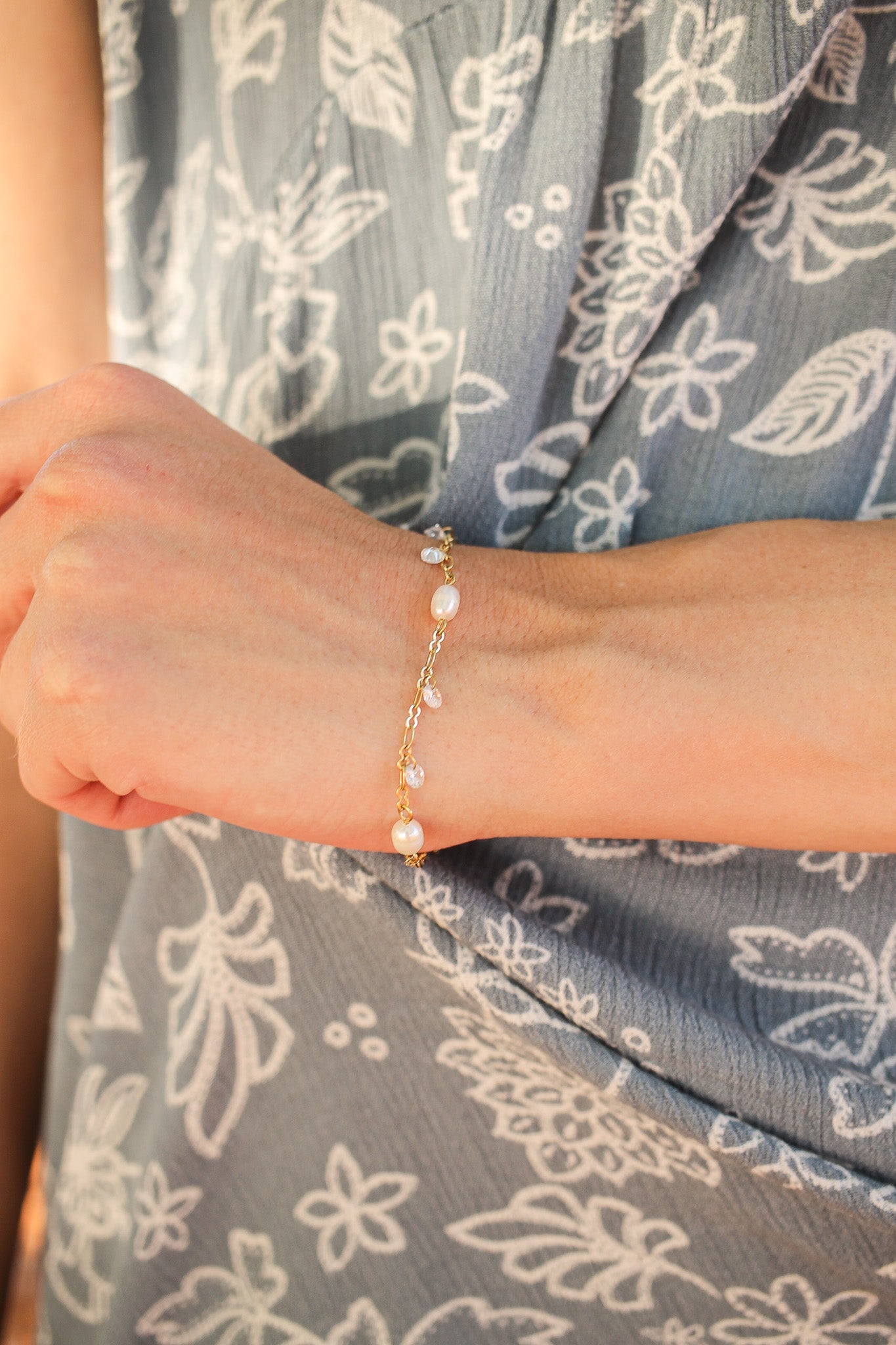 A delicate gold bracelet adorned with small pearls and gemstones rests on a wrist, while the background features a soft fabric with a floral pattern in shades of blue and white.