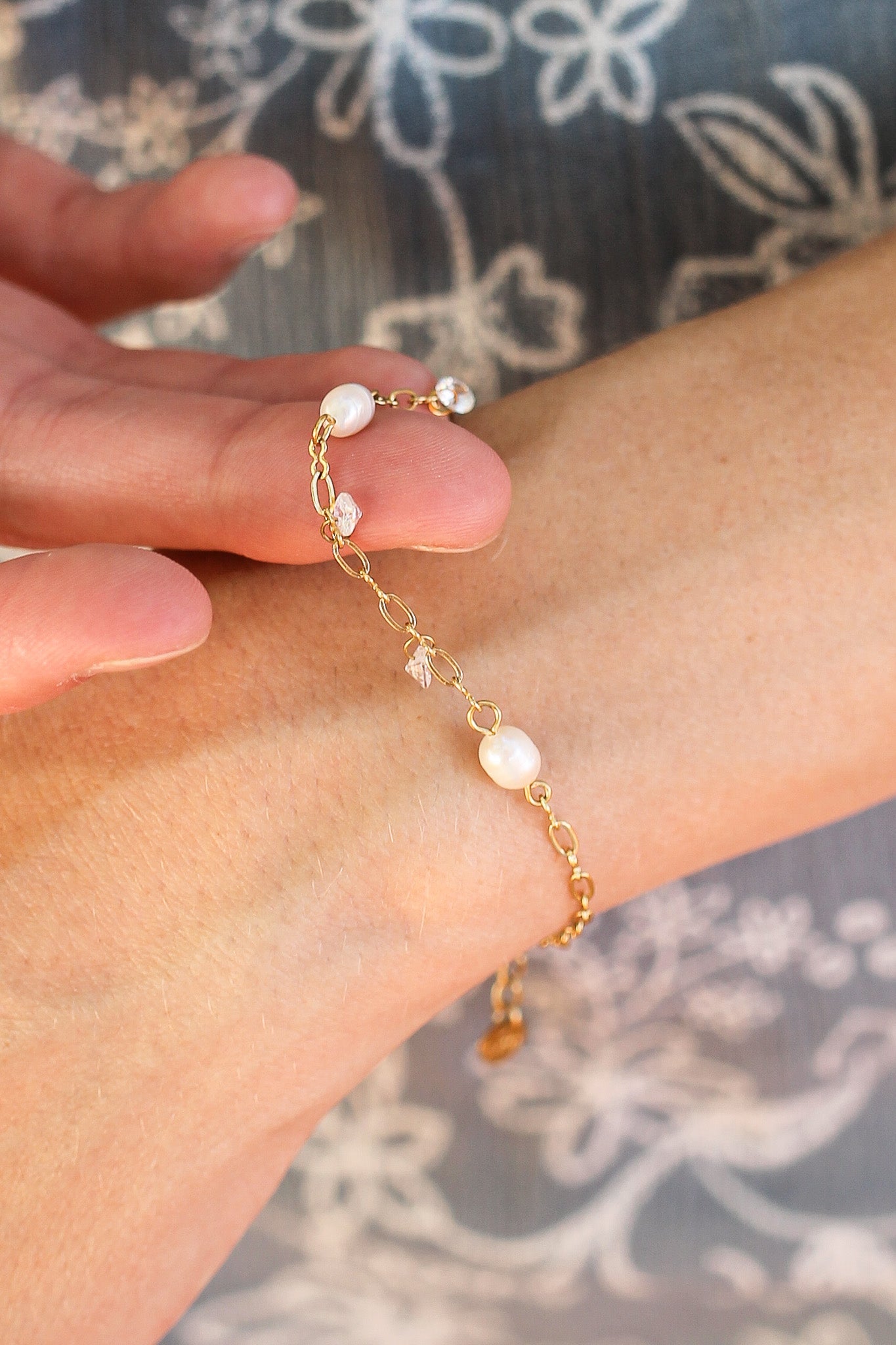A delicate gold bracelet adorned with pearls and crystals is being fastened around a wrist, set against a soft floral-patterned background. The scene conveys elegance and care.